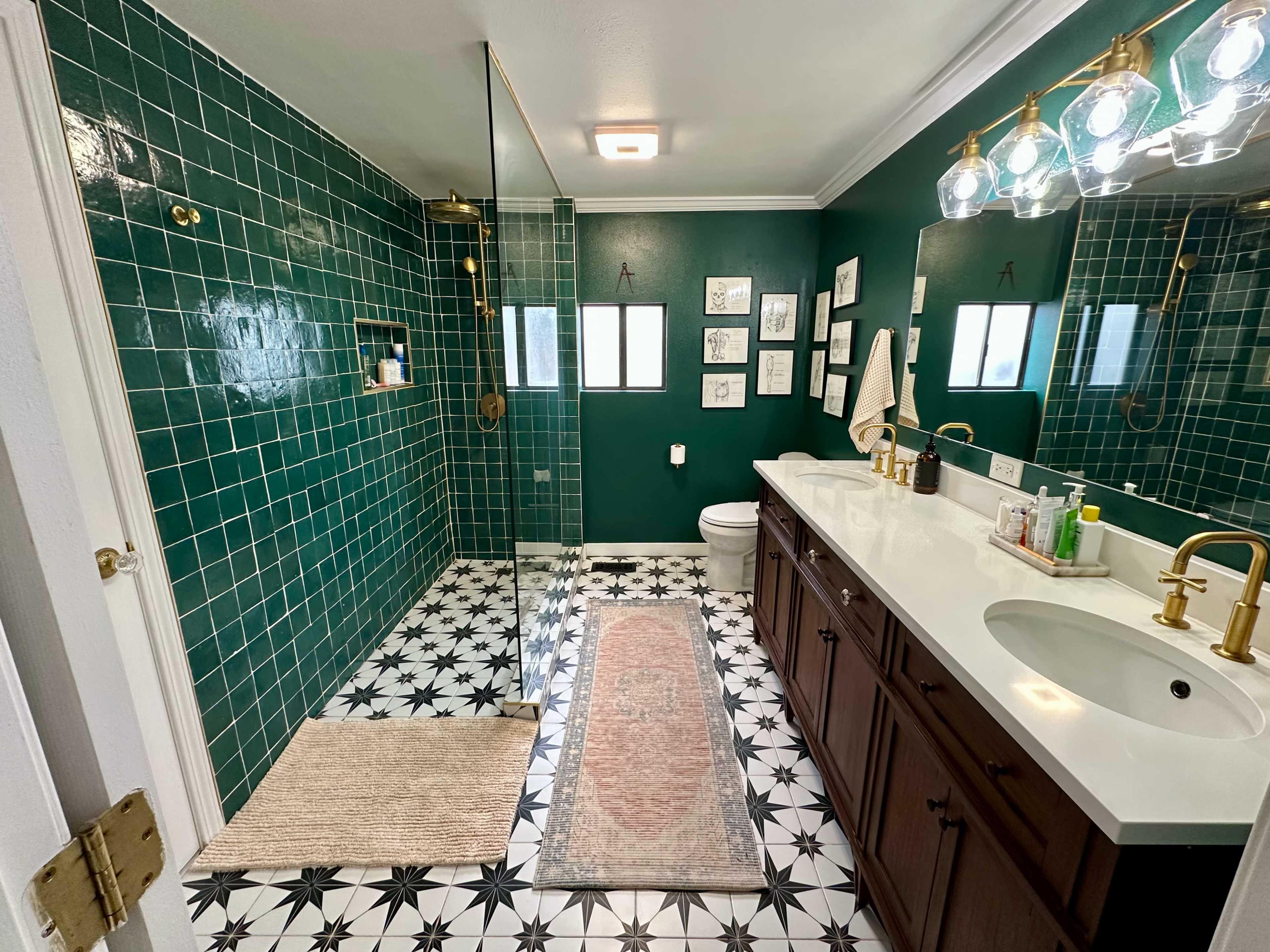 The bathroom features green tiled walls, a glass shower enclosure, a double sink vanity with brass fixtures, and patterned black-and-white floor tiles.