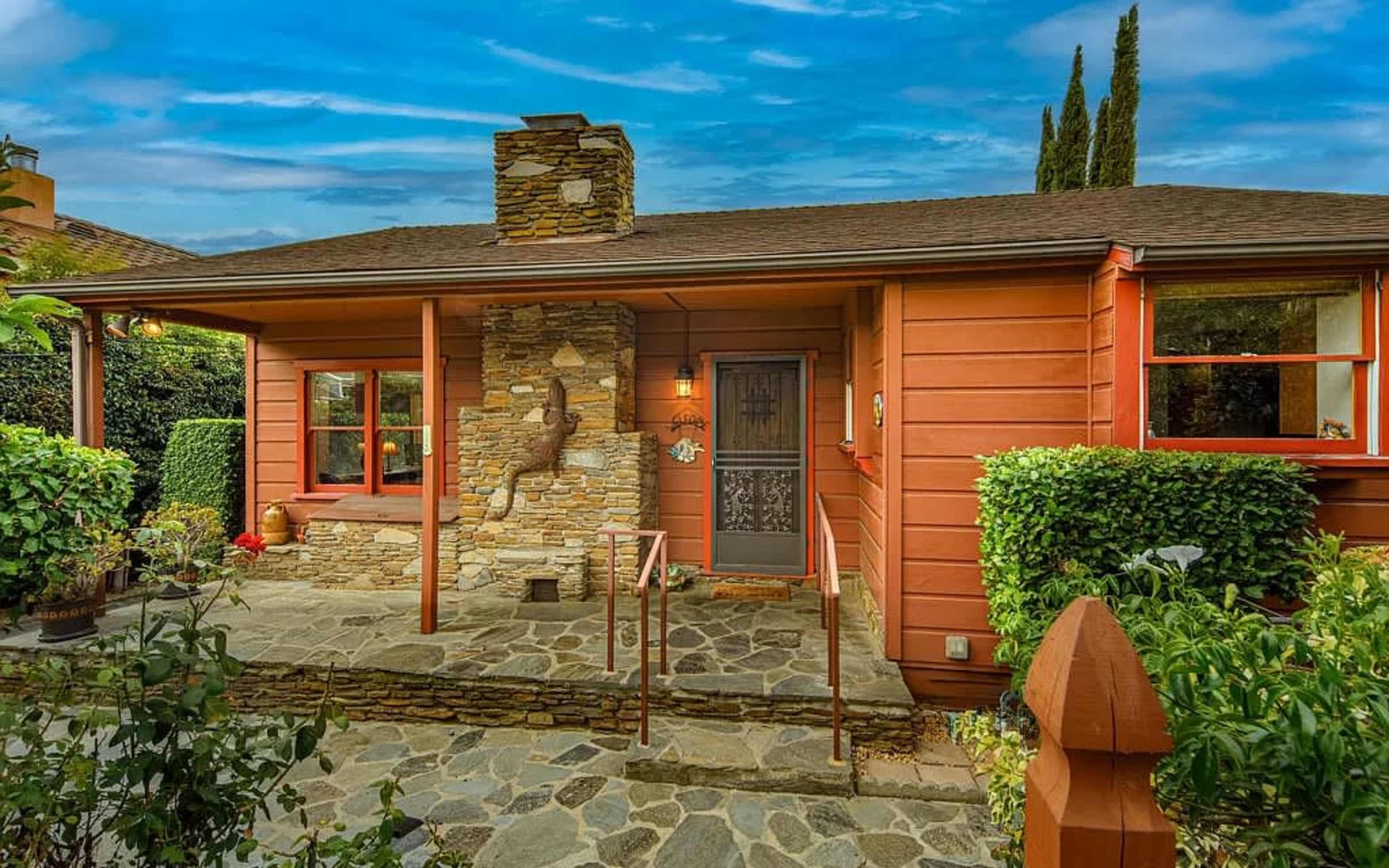 A single-story wooden house with red siding, a stone chimney, and a stone pathway leading to the front door, surrounded by greenery.