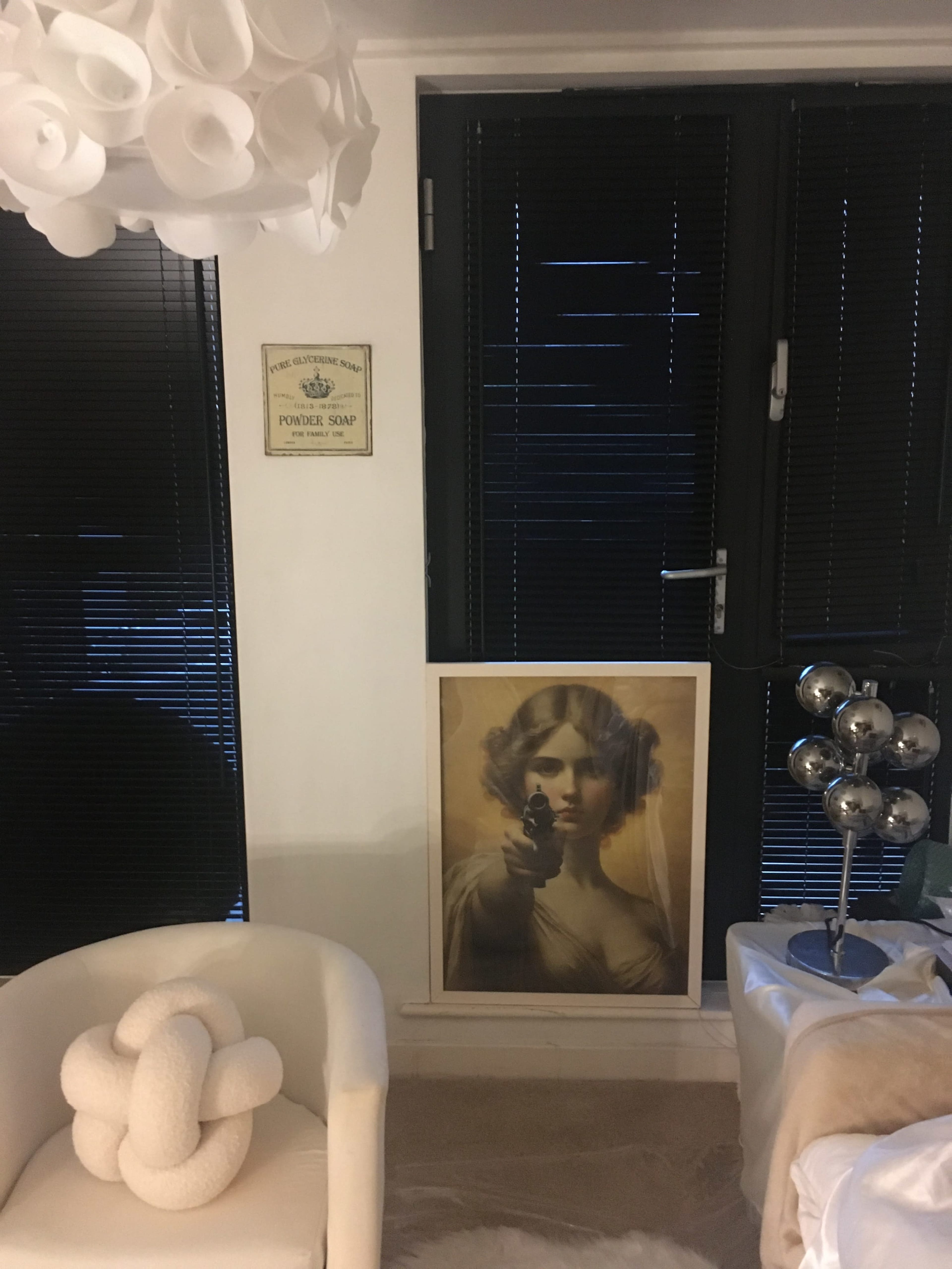 The image shows a room with a modern lamp, a poster of a woman holding a gun, and black blinds on the window.