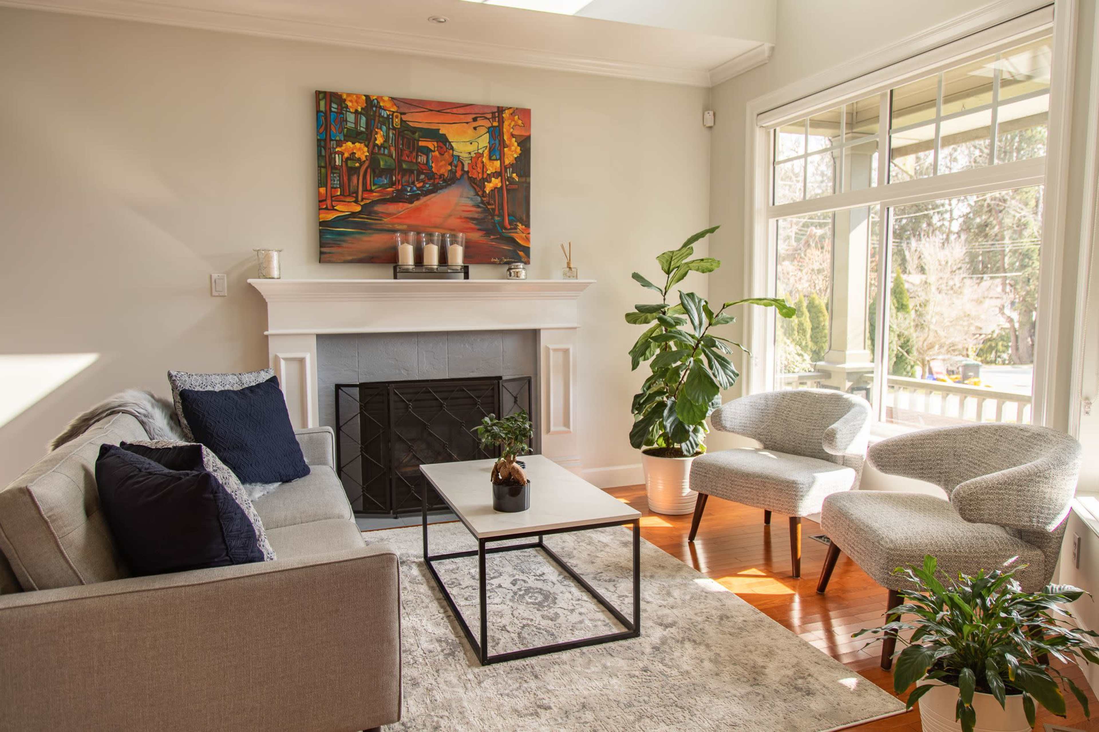 A modern living room features a gray sofa, two armchairs, a coffee table, and a fireplace, complemented by large windows and a colorful painting on the wall.