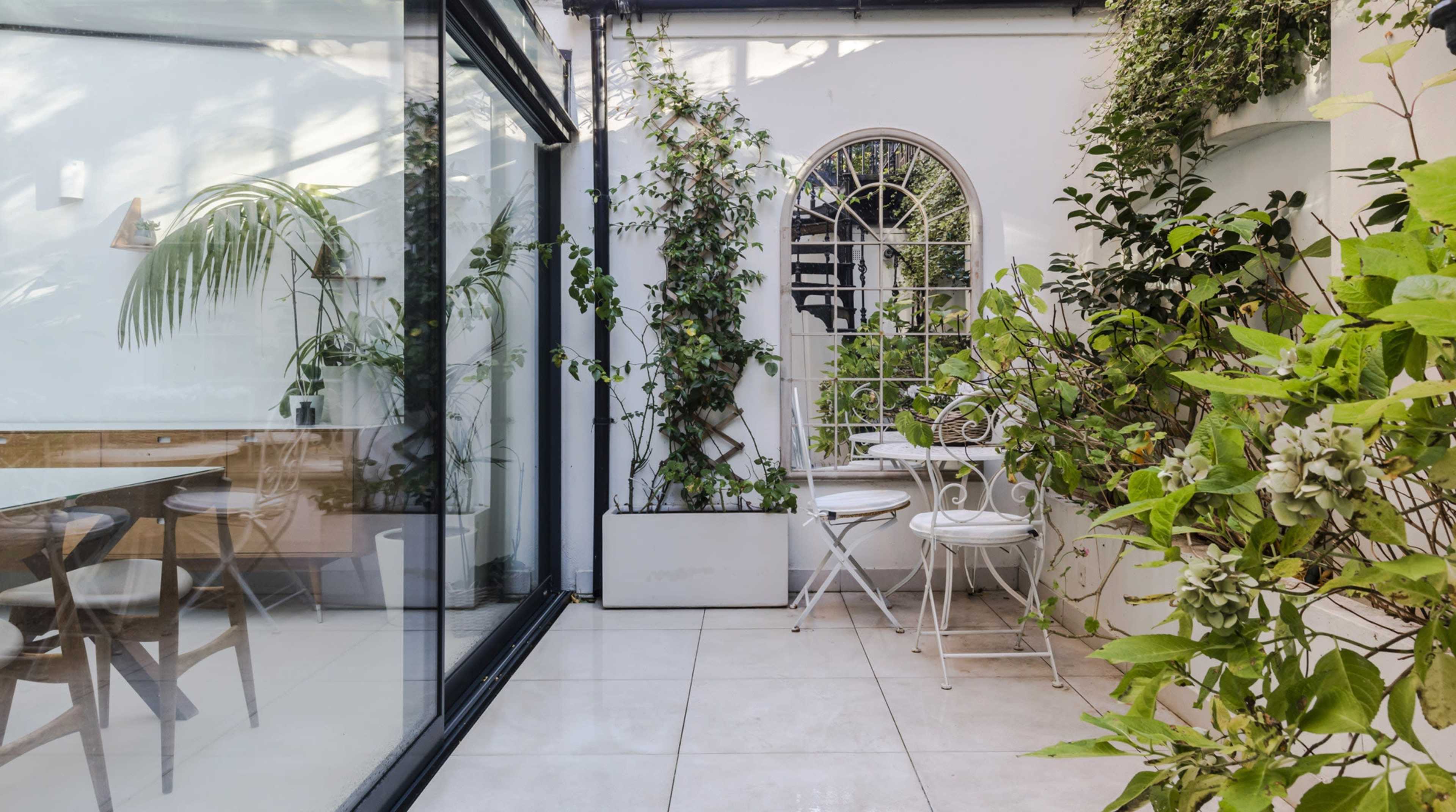 A small indoor garden area enclosed by glass walls, featuring plants, a white metal table, and chairs.