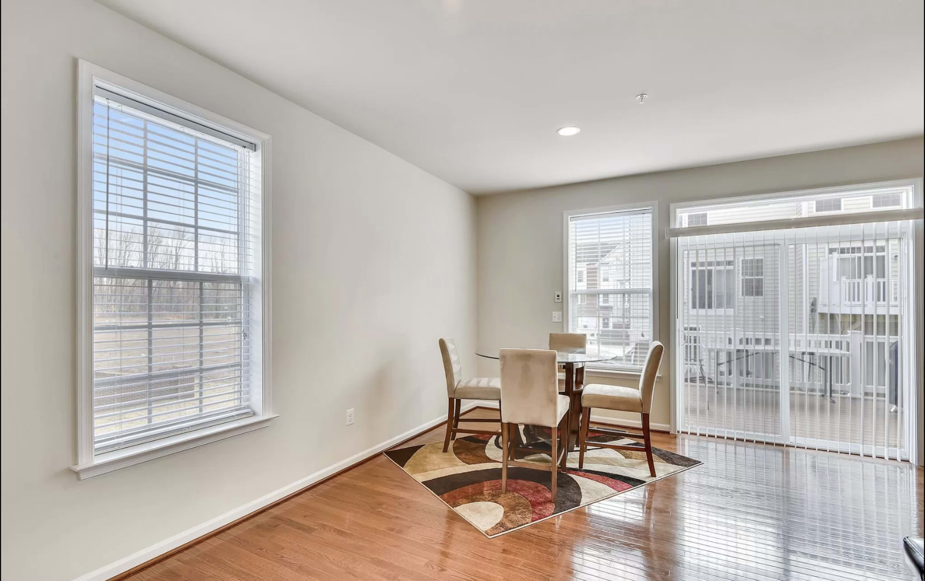 A dining area with a circular table and four chairs is adjacent to large windows, featuring a hardwood floor and a patterned area rug.