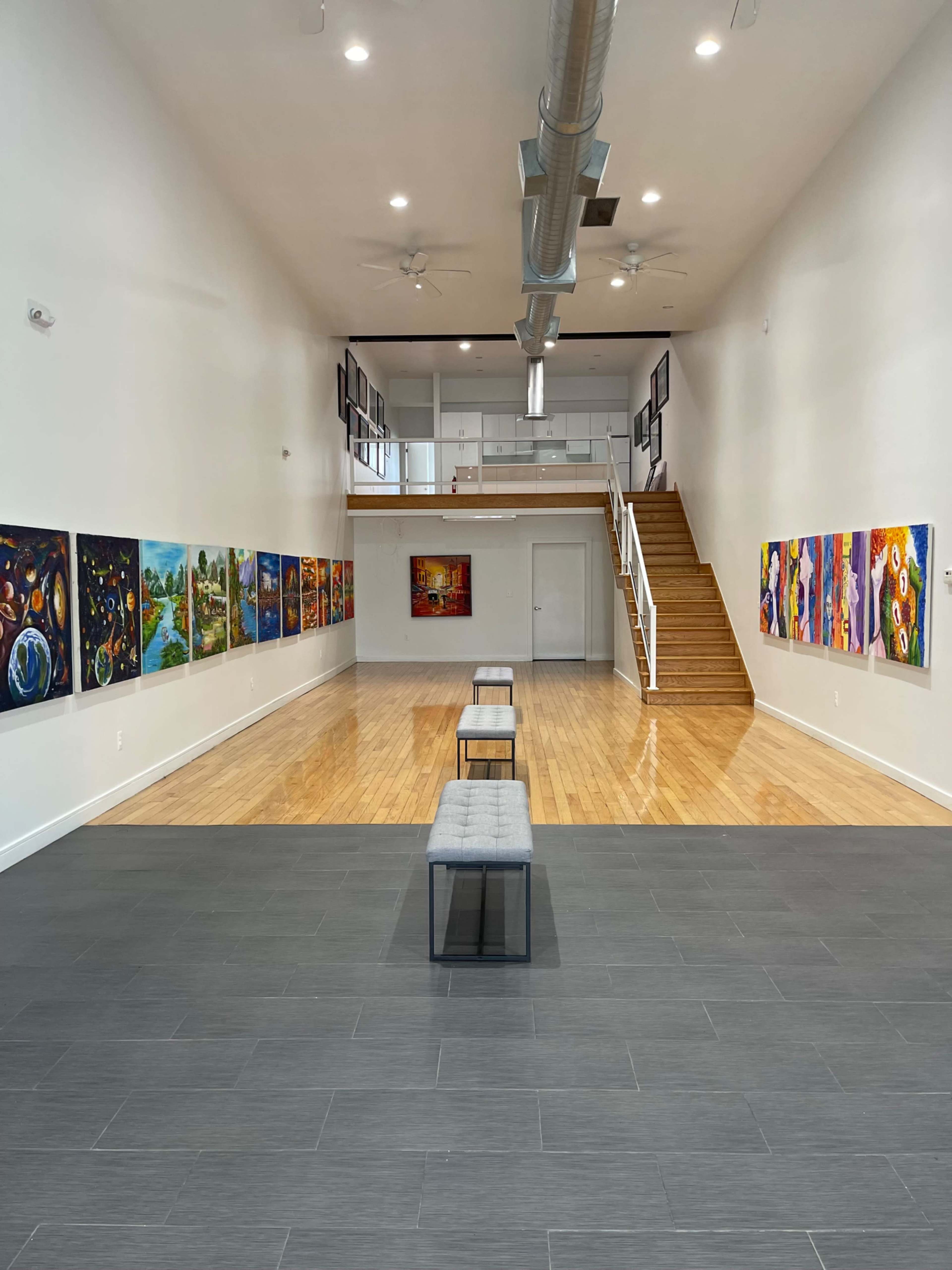 A spacious art gallery features colorful paintings on the walls with wooden floors and a staircase at the end of the room.