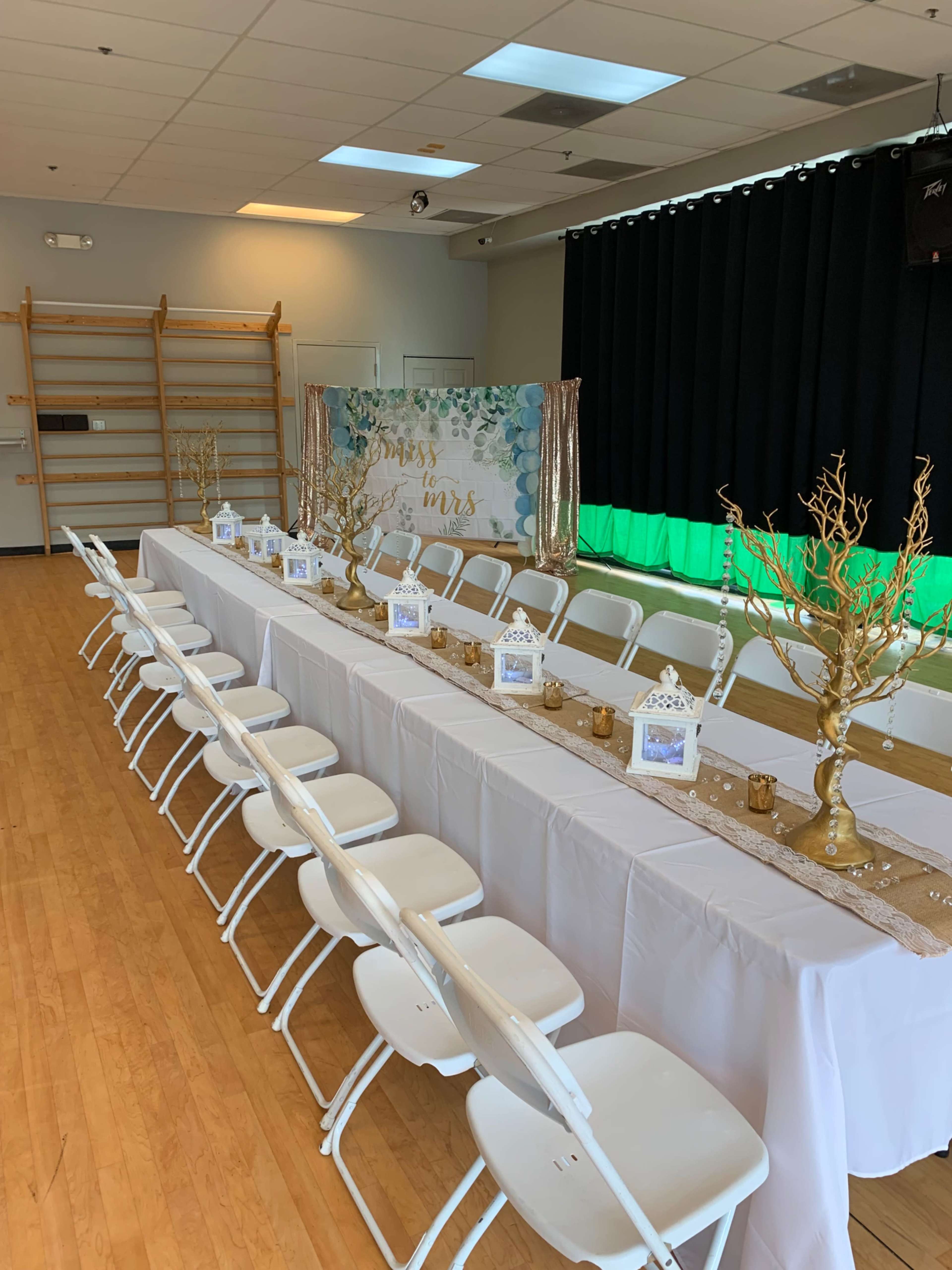 The image shows a long banquet table set up in a spacious room with white chairs and decorative centerpieces, including gold-painted branches and small lanterns.