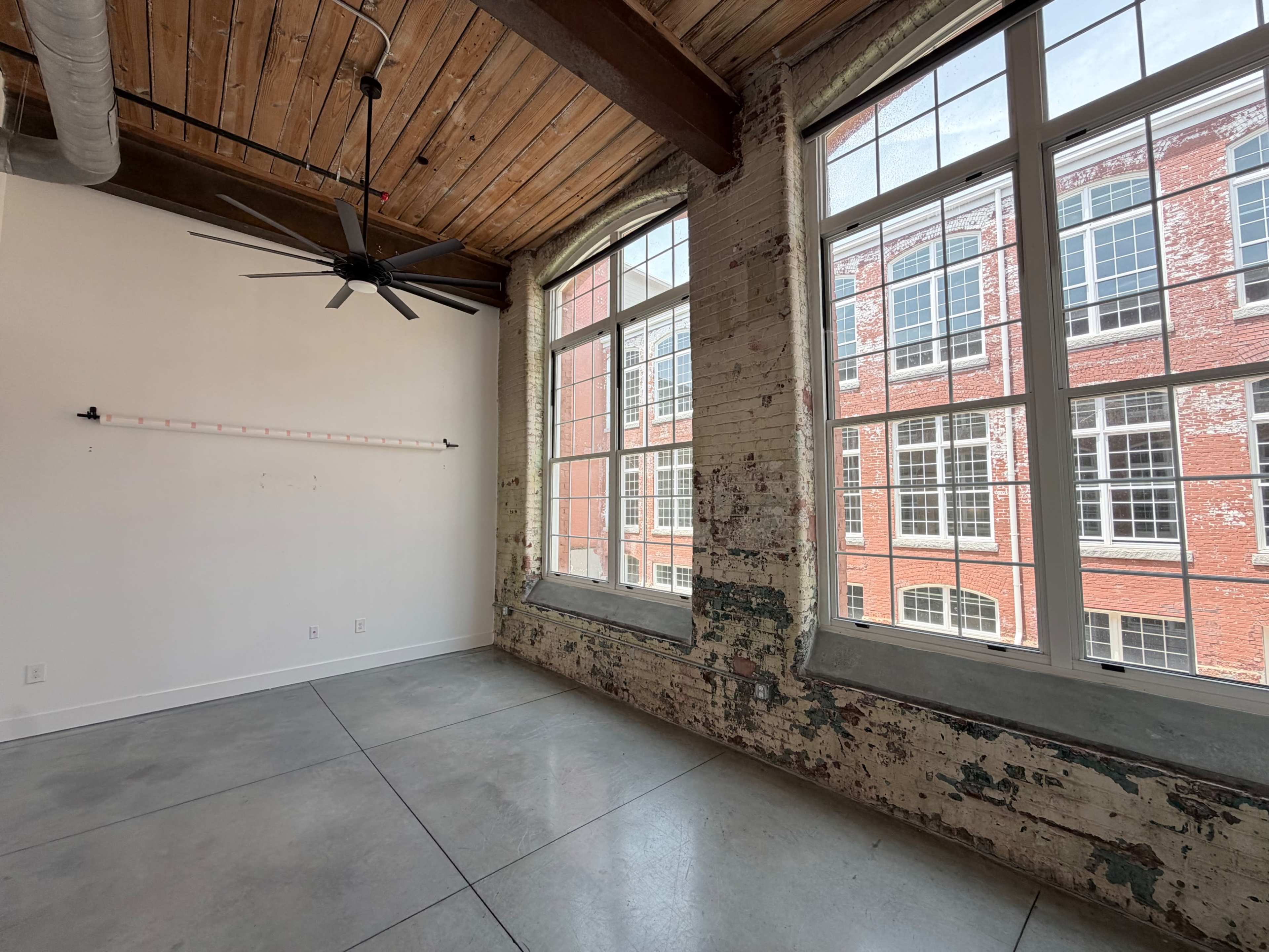 The image shows an empty room with a concrete floor, exposed brick walls, large windows, and wooden ceiling beams.