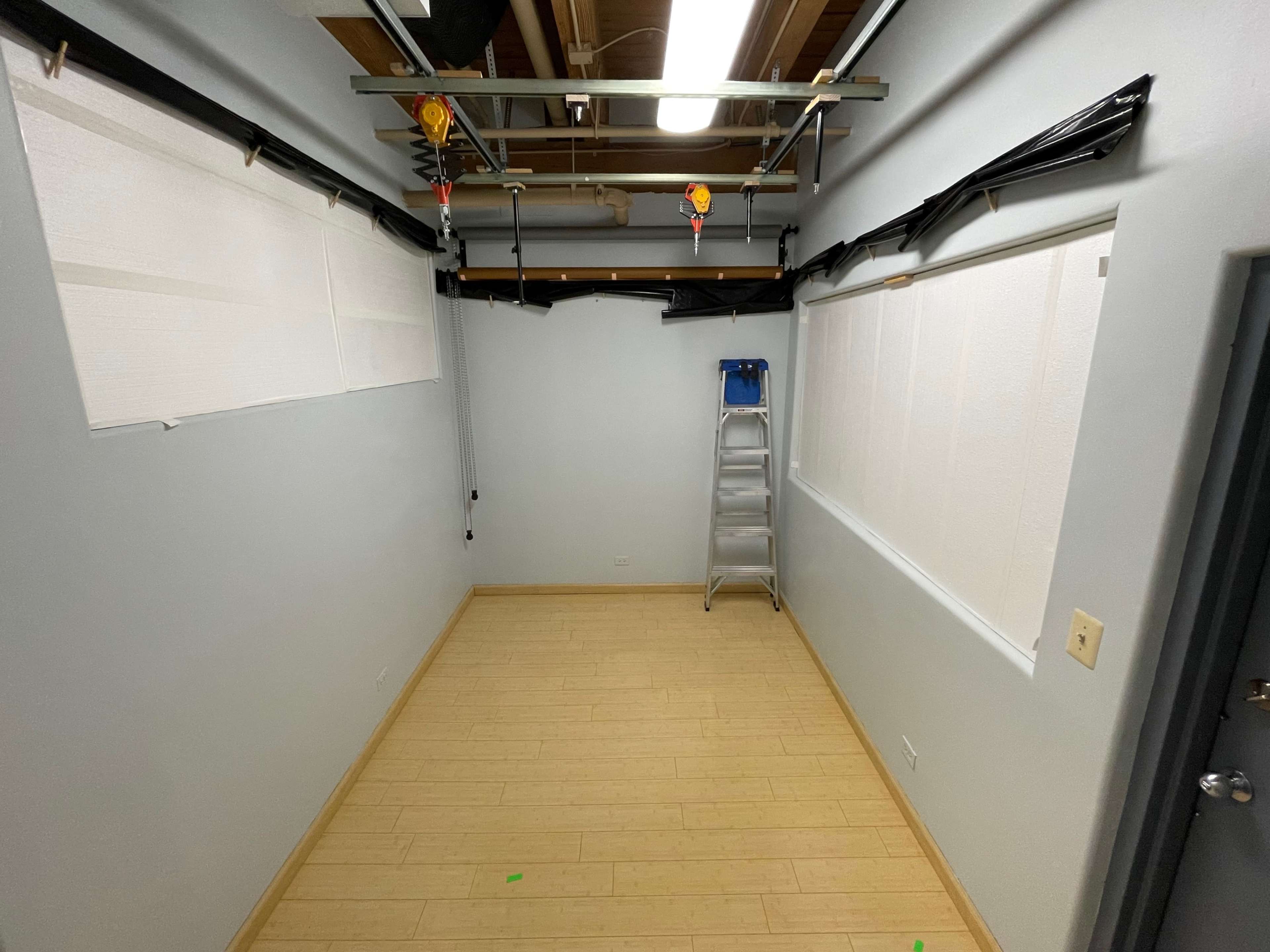 A vacant room with light-colored walls, a ladder against one wall, and unadorned windows covered with white blinds.