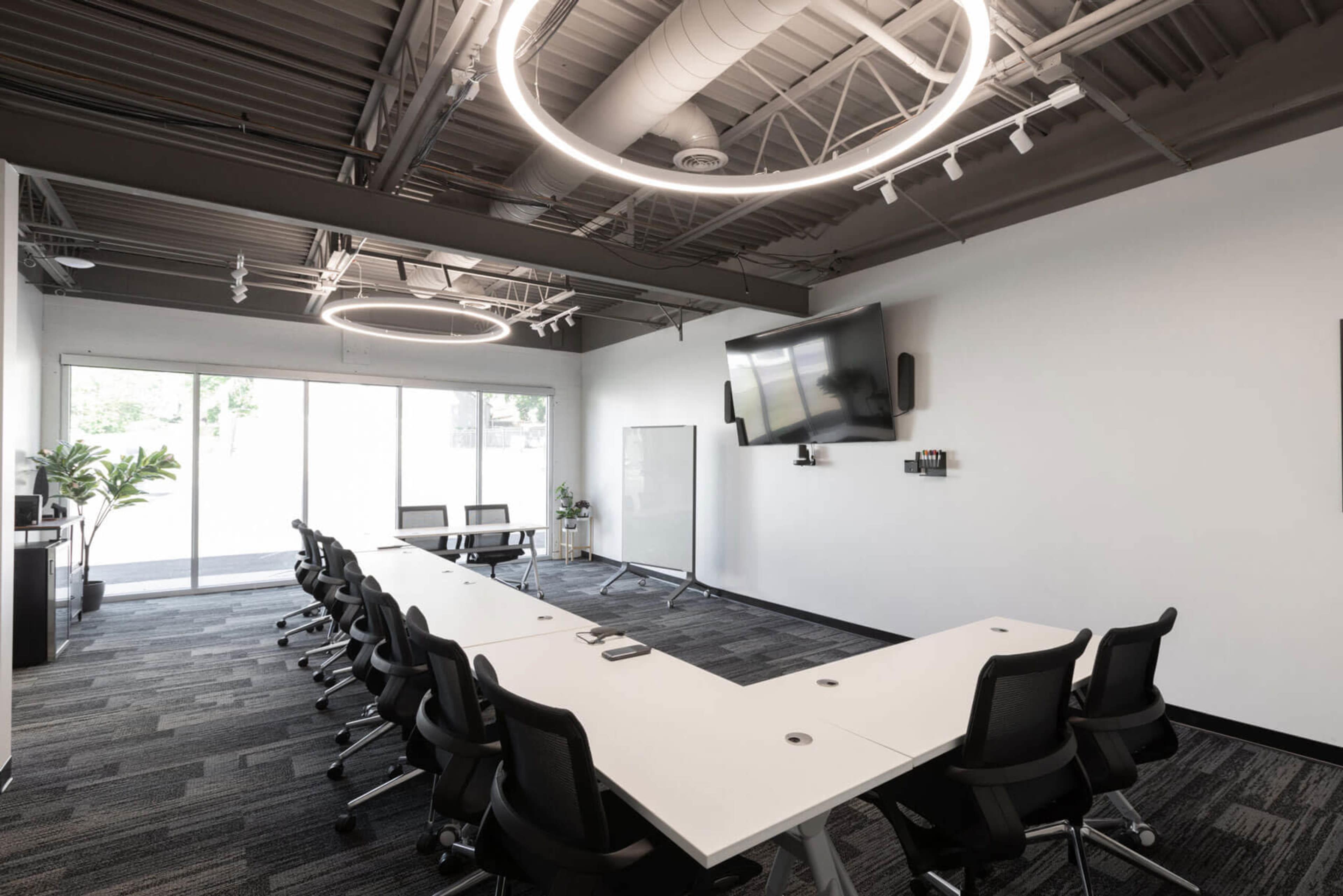 A modern conference room with a long, white table surrounded by black chairs, large windows, and circular light fixtures overhead.