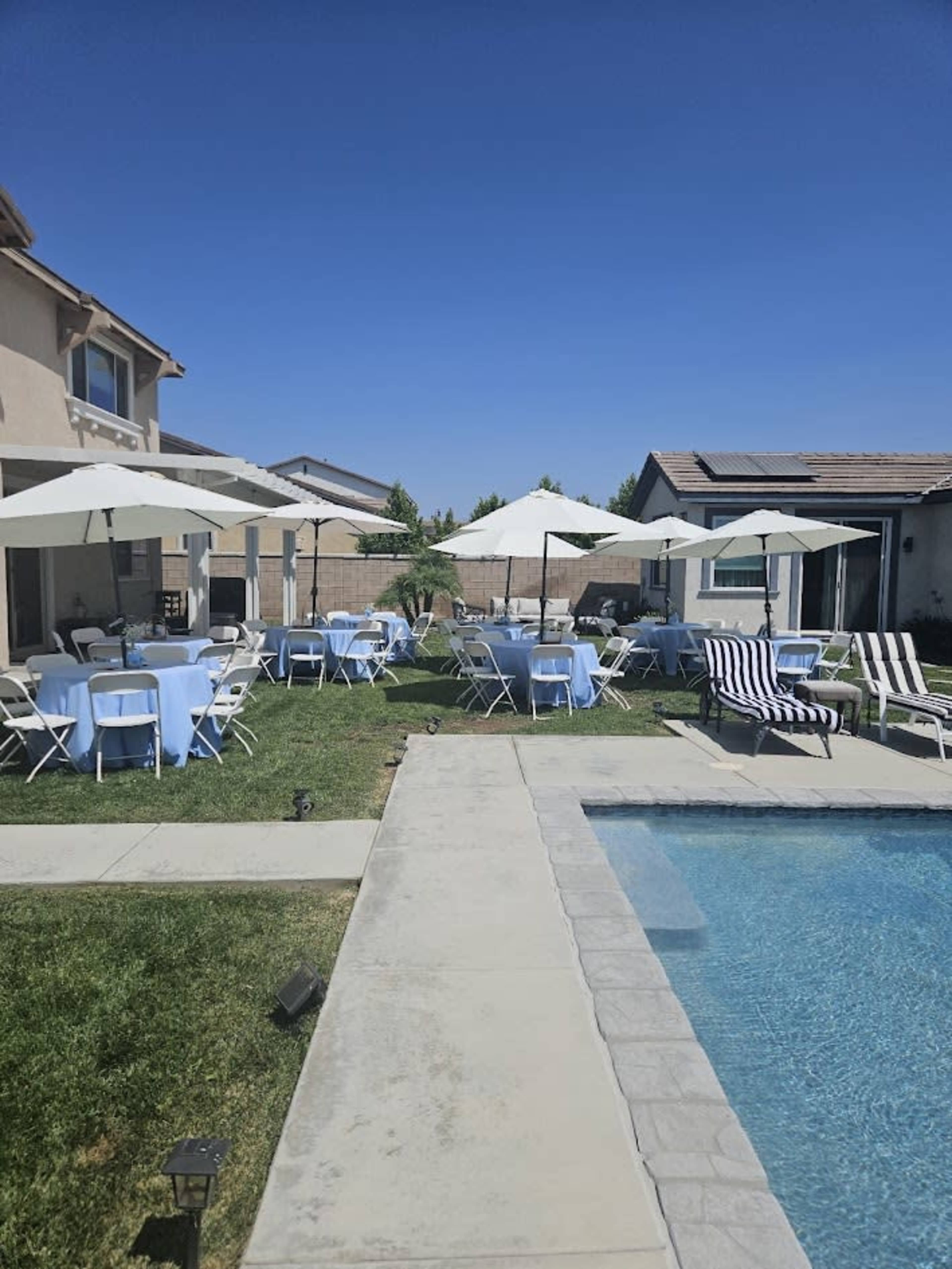 A backyard scene features several tables with light blue tablecloths, white chairs, and large umbrellas, all set up near a swimming pool.