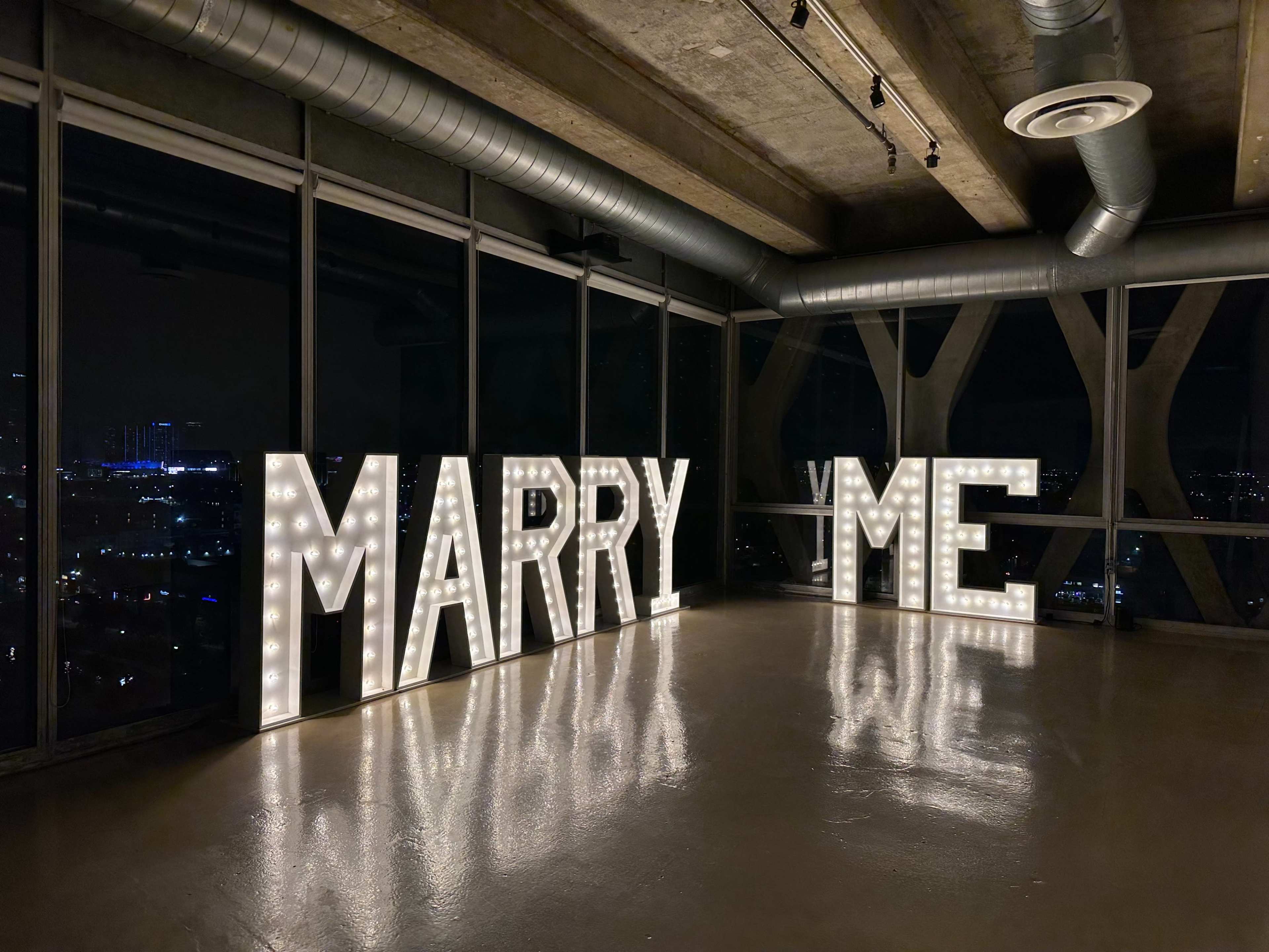 The scene features large, illuminated letters spelling "MARRY ME" in a dimly lit room with city lights visible through windows.