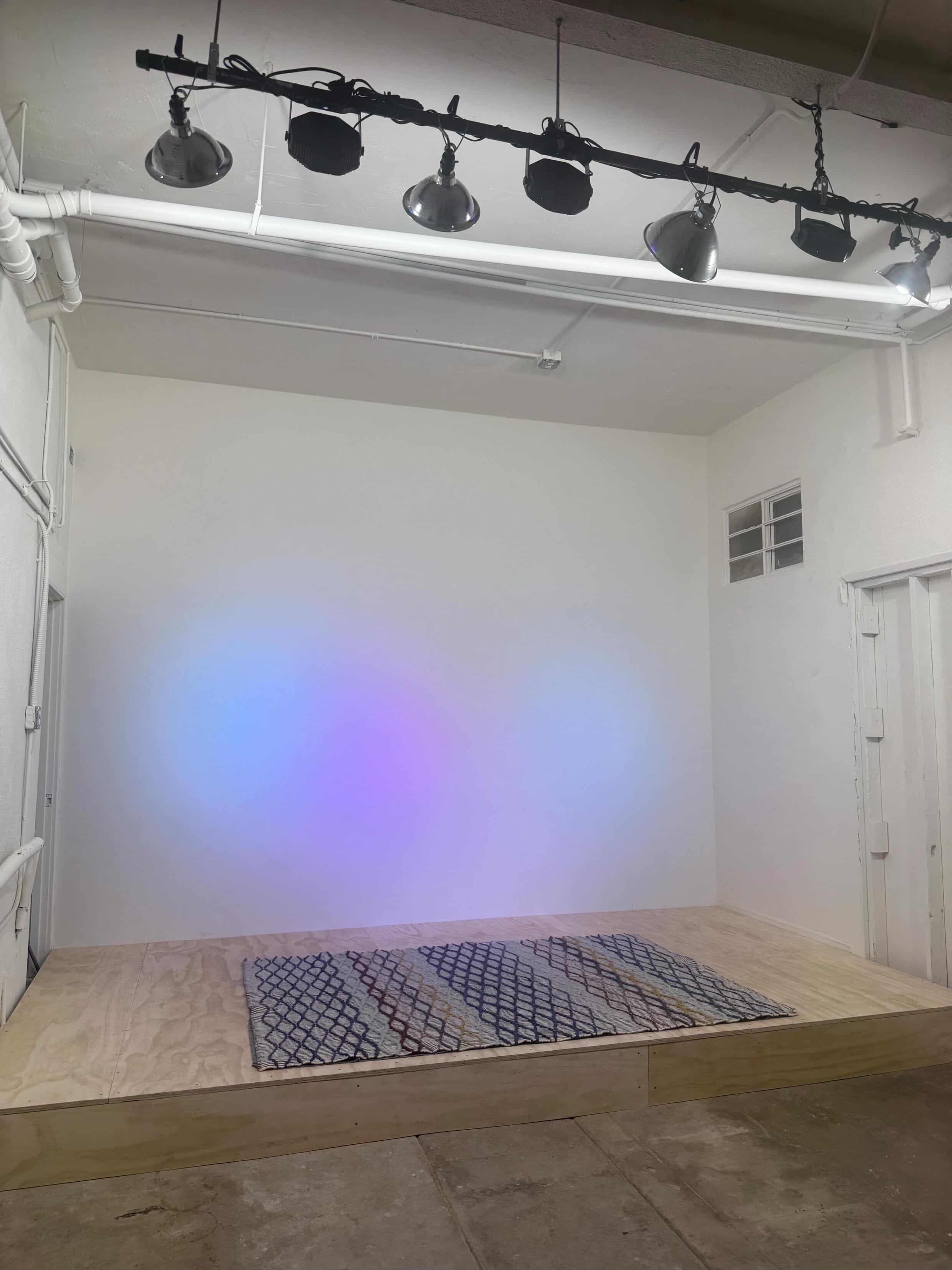 The image shows an empty stage area with a wooden platform, overhead spotlights, and a wall displaying soft, colorful lighting.