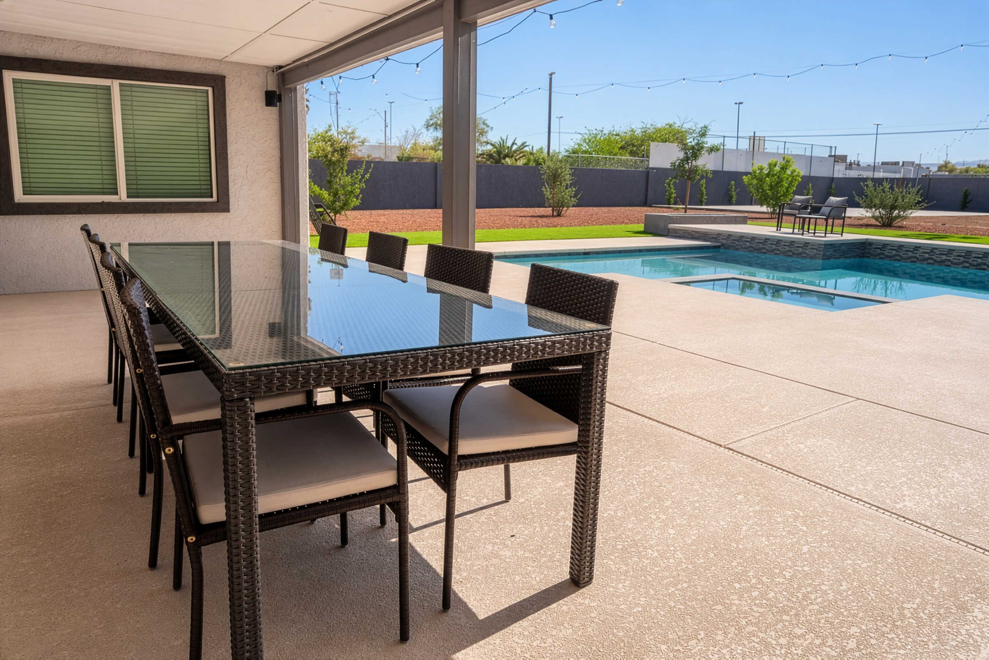 A modern patio features a glass dining table with eight chairs beside a swimming pool and landscaped greenery.