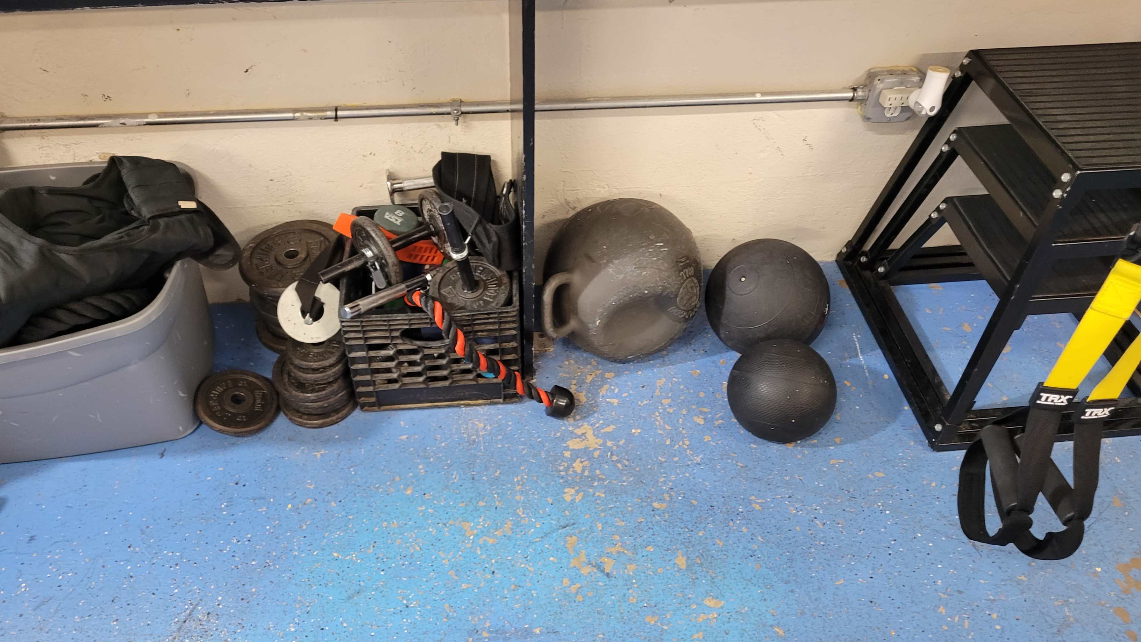 The image shows a workout space with various exercise equipment, including weights, a kettlebell, and a suspension trainer, arranged near a storage container.