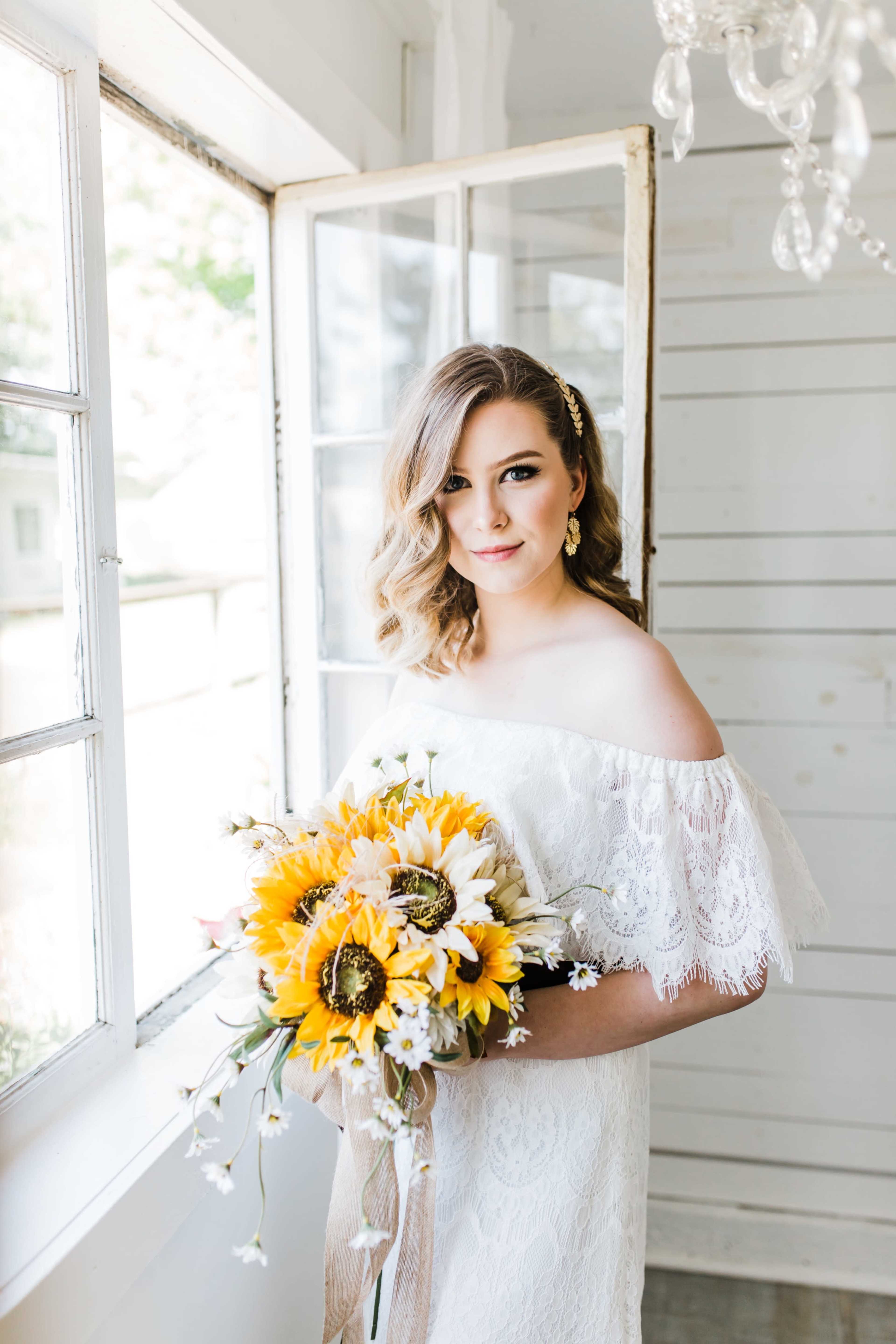 A woman in a white off-the-shoulder dress holds a bouquet of sunflowers and other flowers while standing by an open window.