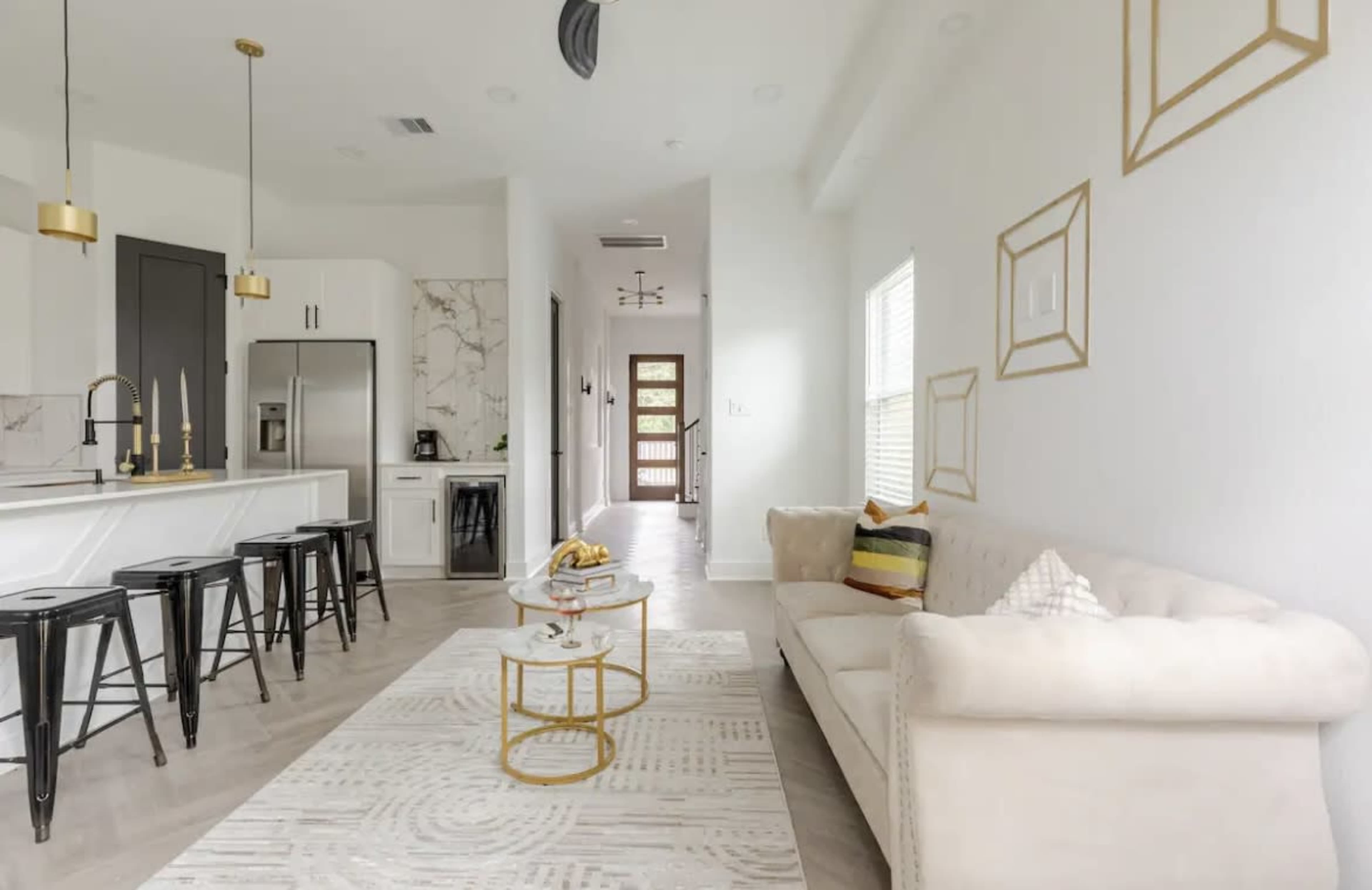 A modern living space featuring a light-colored sofa, a coffee table, a patterned rug, and a kitchen area with bar stools.