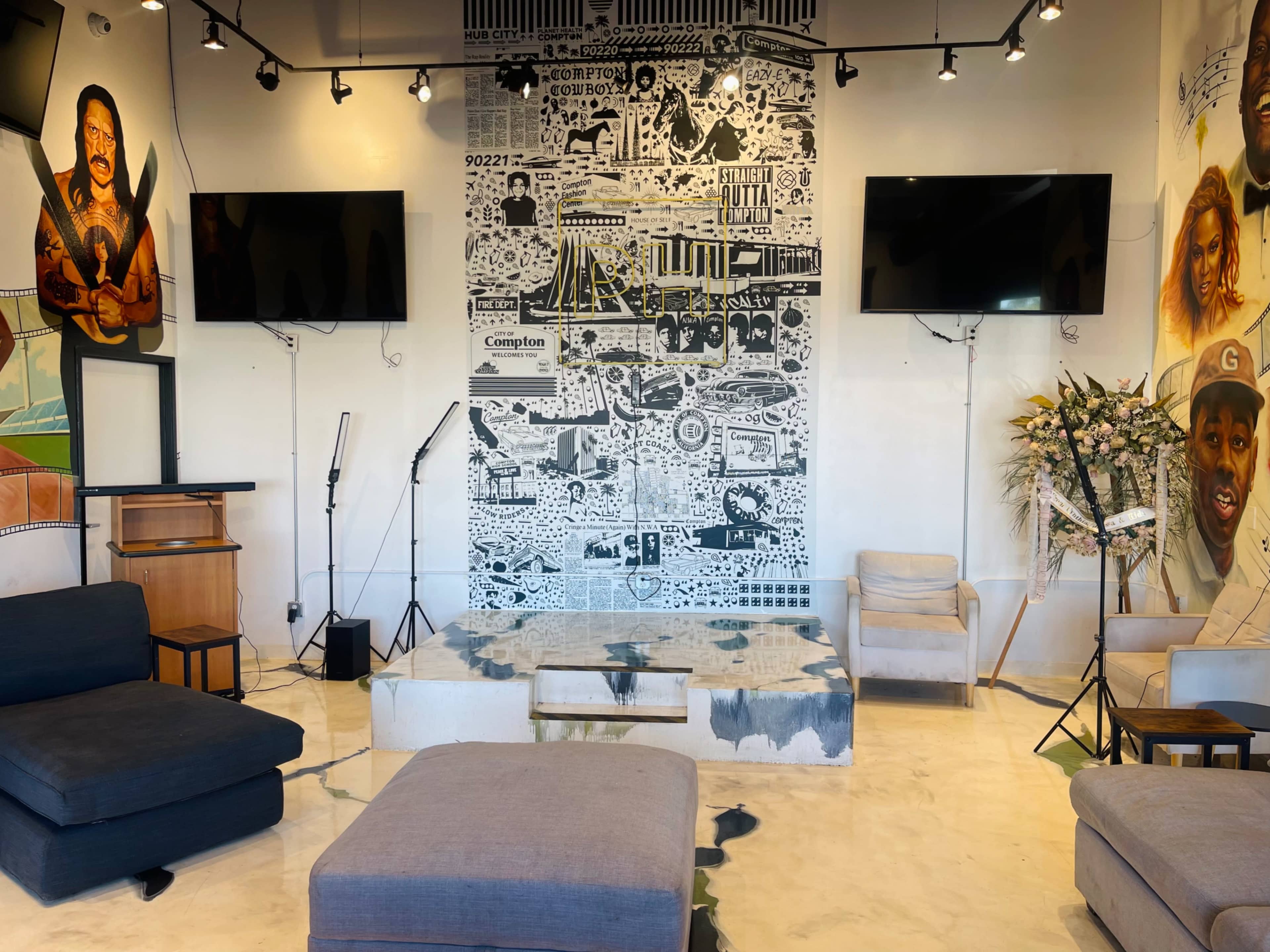 The scene shows a modern, minimalist lounge area featuring gray seating, a mural-covered wall, and two wall-mounted televisions.