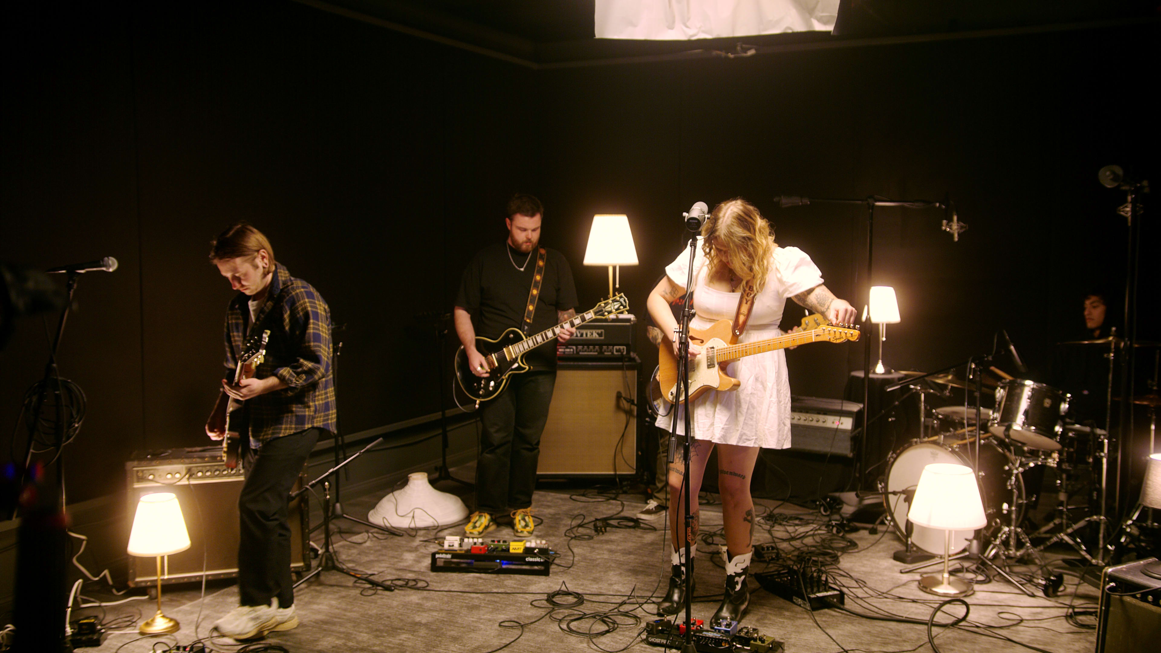 A band performs on stage in a dimly lit studio, surrounded by several lamps and various instruments.