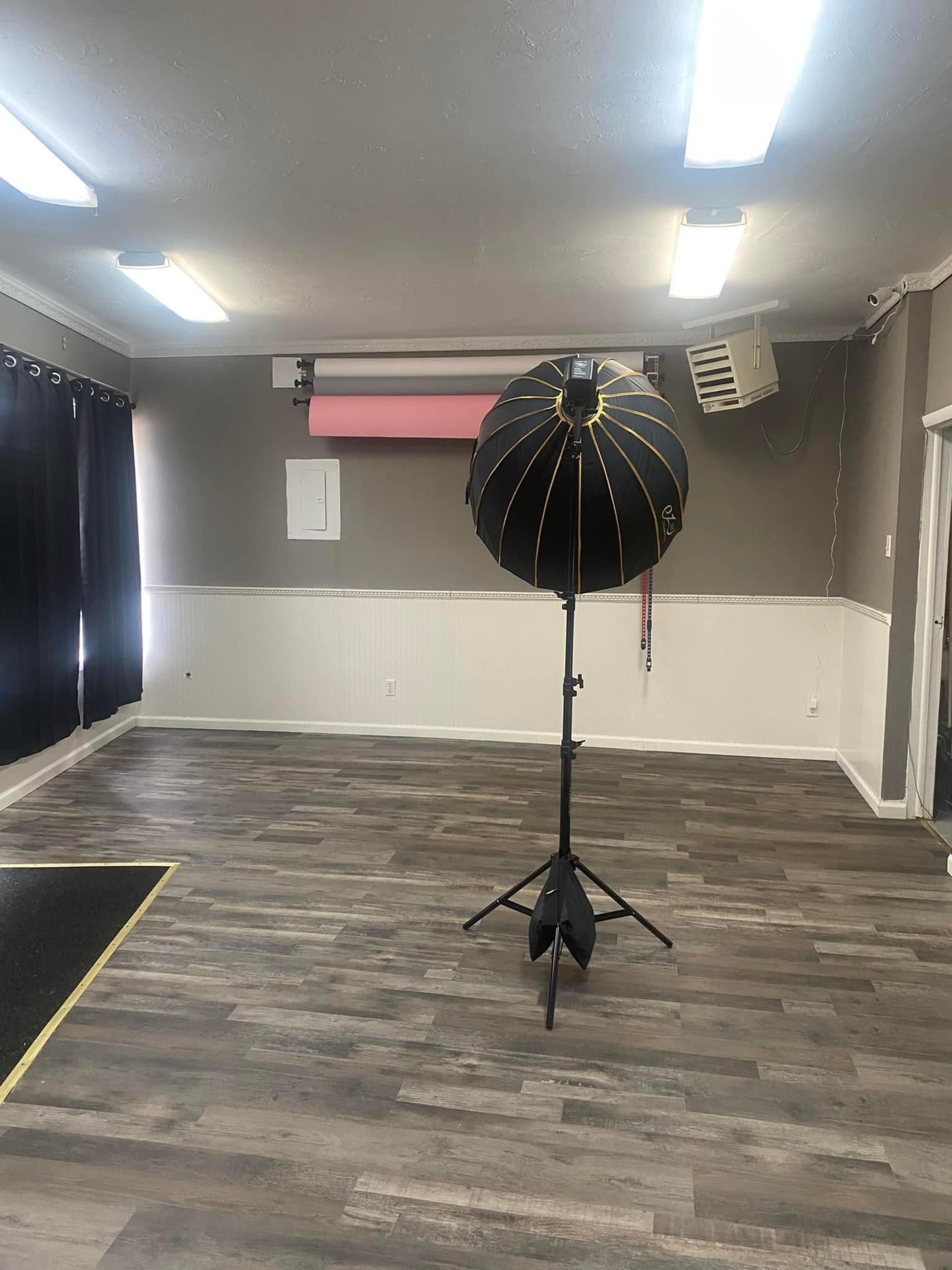 An empty room with wooden flooring, featuring a large photography light umbrella on a stand, dark curtains on one side, and a backdrop holder with a pink backdrop hanging on the wall.