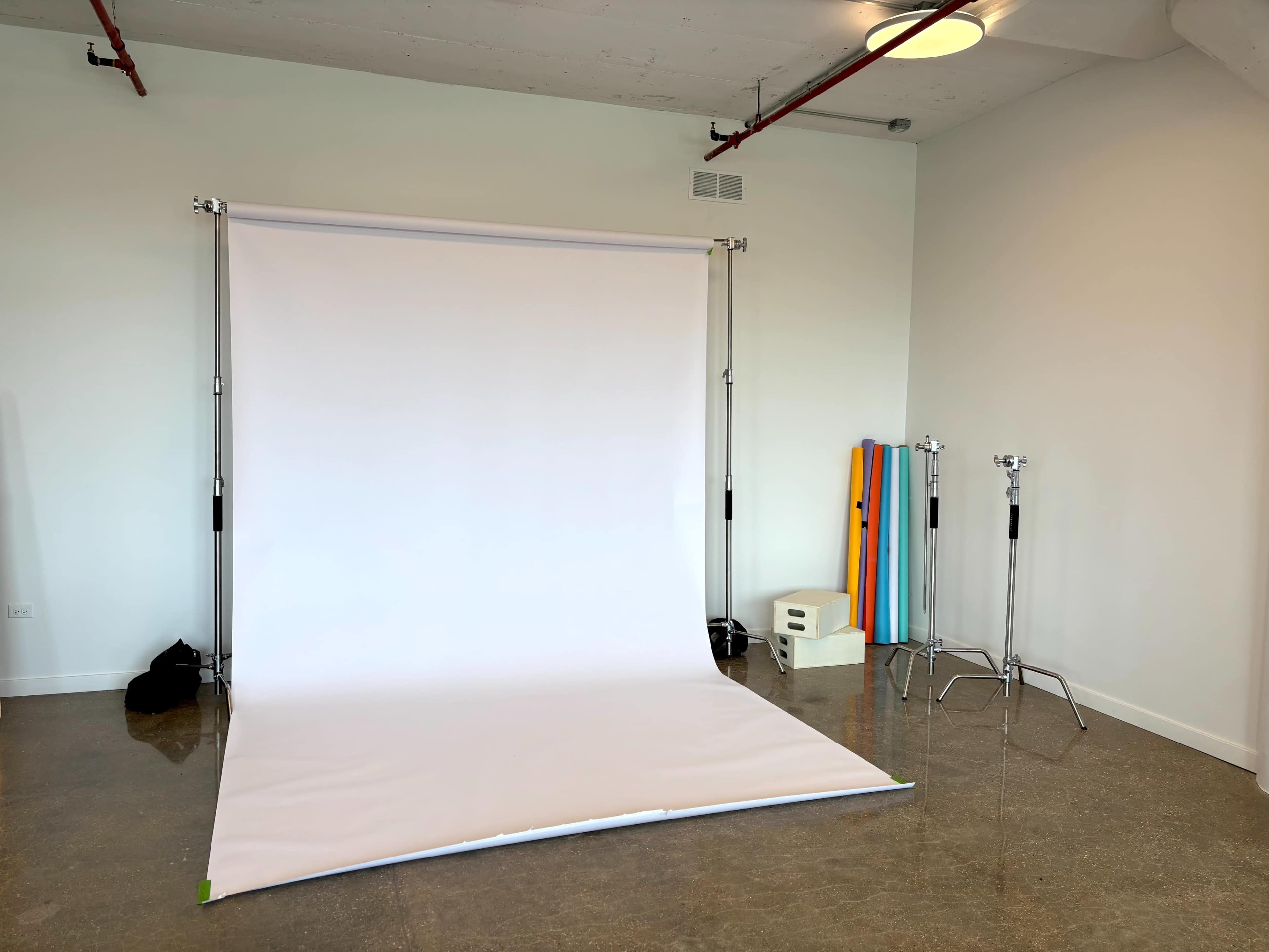 The image shows a photography studio with a white backdrop setup, two light stands, and colorful backdrop rolls arranged in a corner.