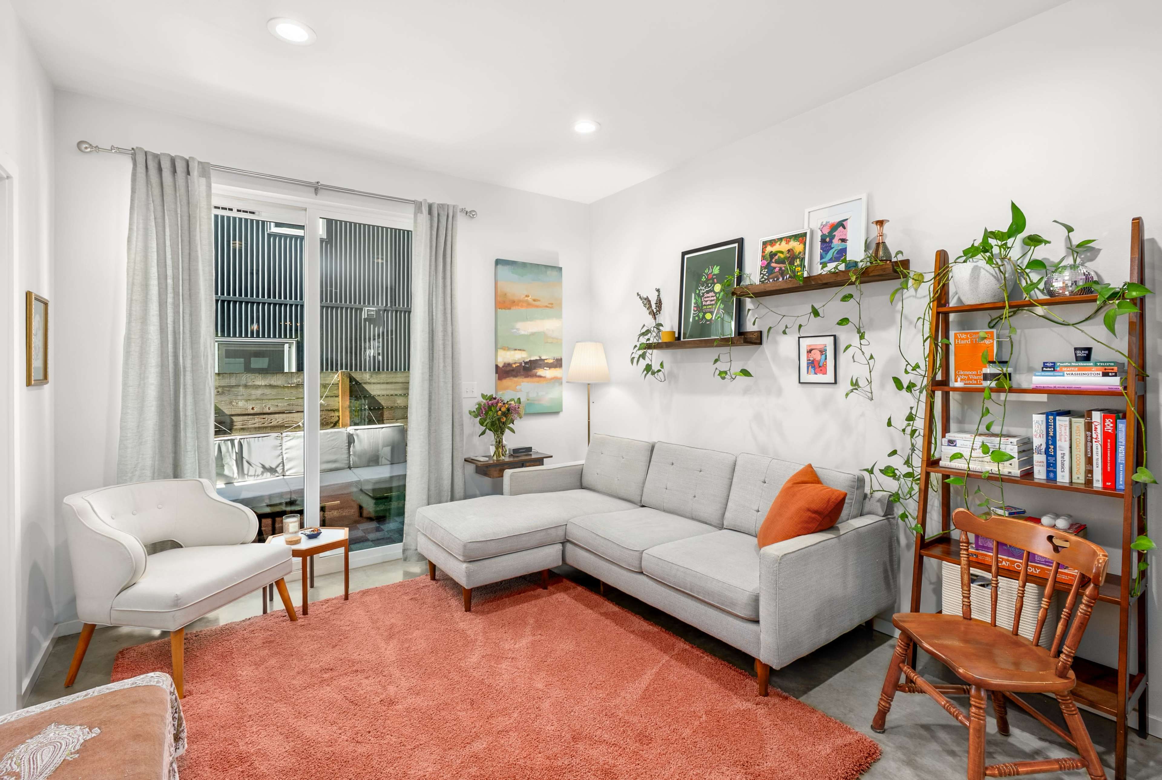 The image shows a modern living room featuring a sectional sofa, a small chair, a bookshelf with plants, and large windows letting in natural light.