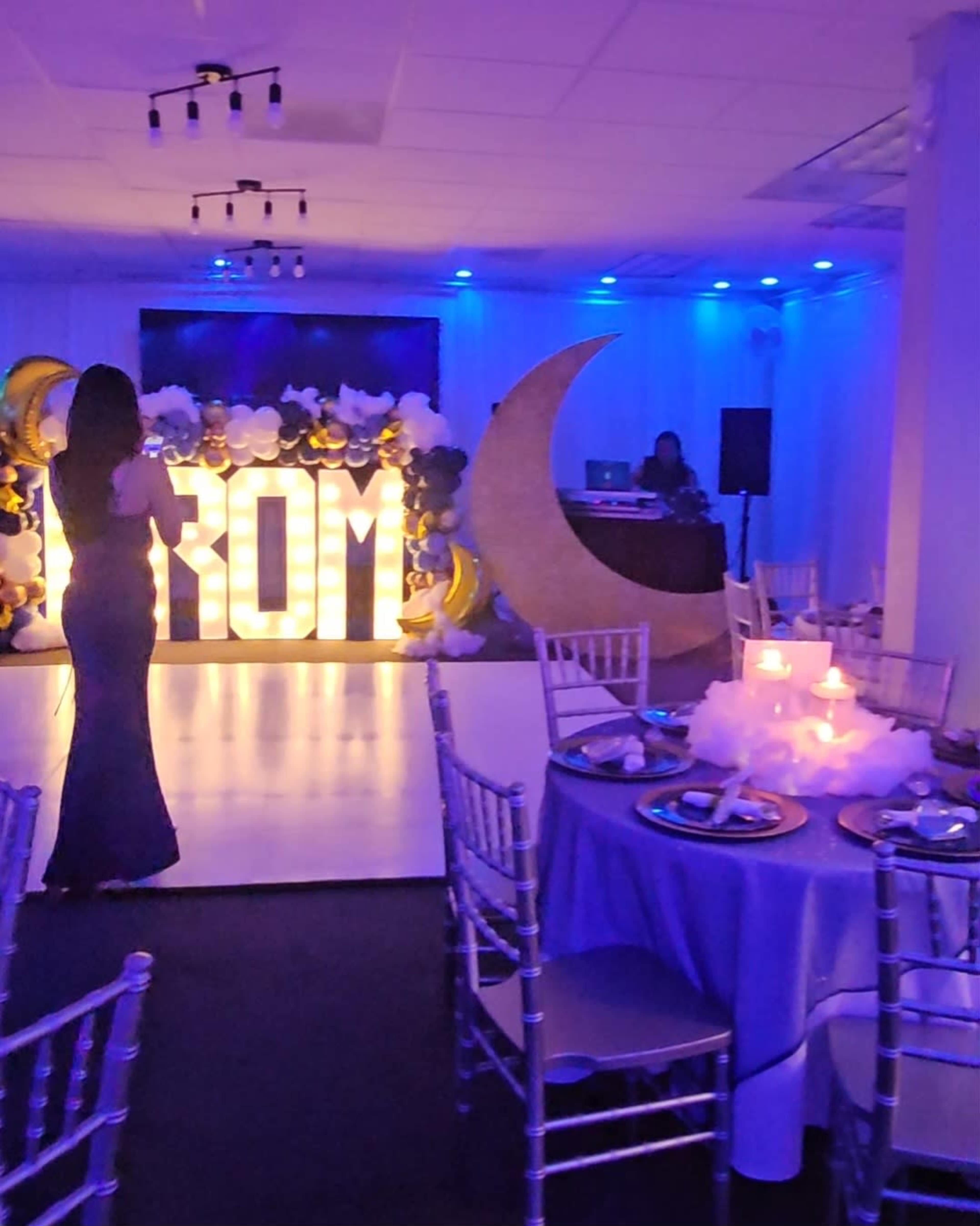 The image shows an event space decorated for a prom, featuring a large illuminated "PROM" sign, table settings with candles, and a backdrop of balloons and a crescent moon.