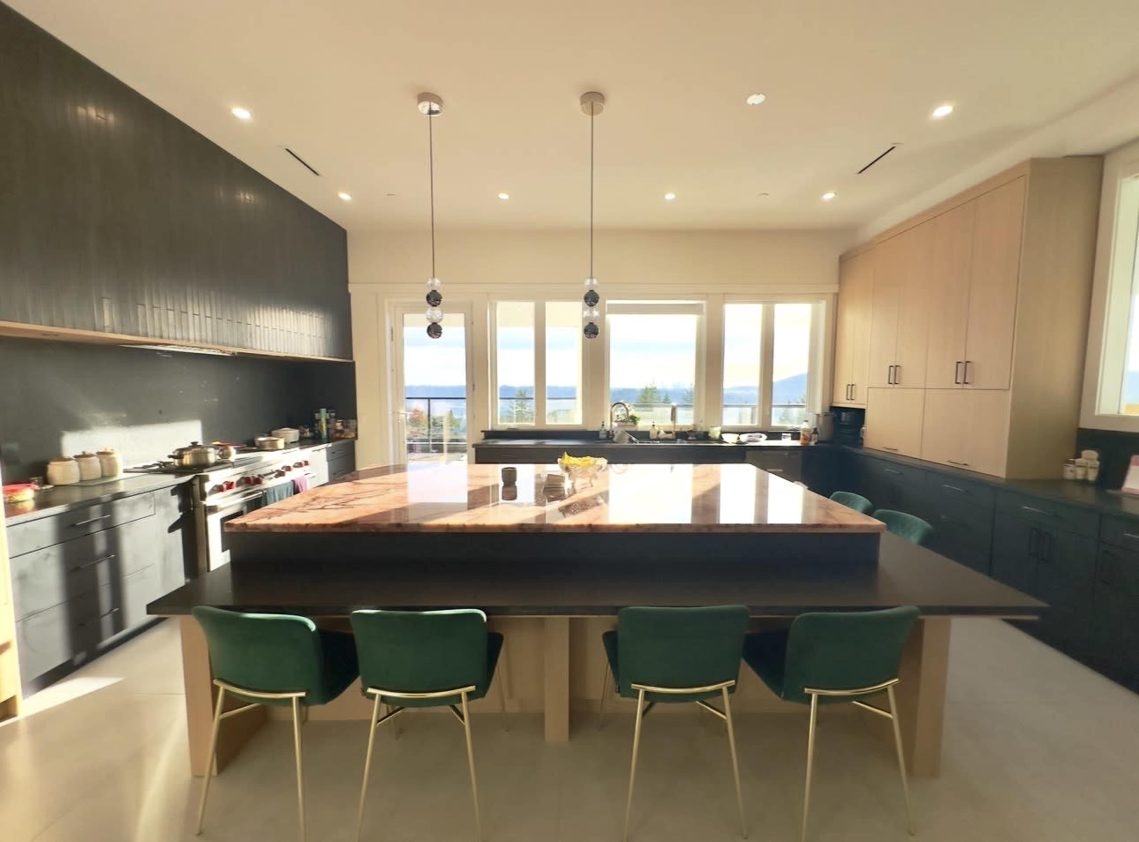 A spacious kitchen features a large island with a dark countertop, green chairs, and large windows that overlook a view of hills and water.