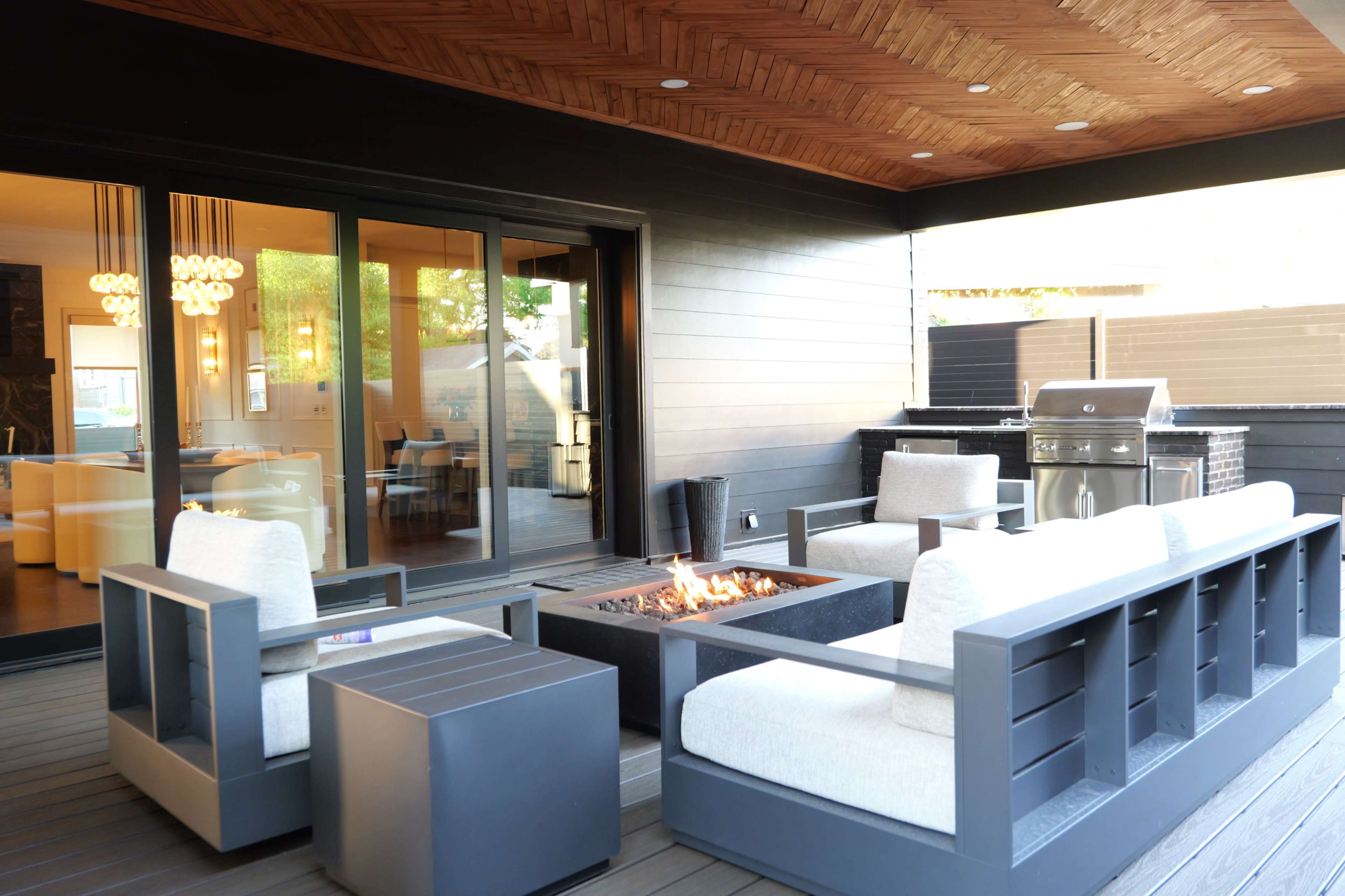 The image shows a modern outdoor seating area featuring gray sofas around a fire pit, with sliding glass doors leading to an indoor space.