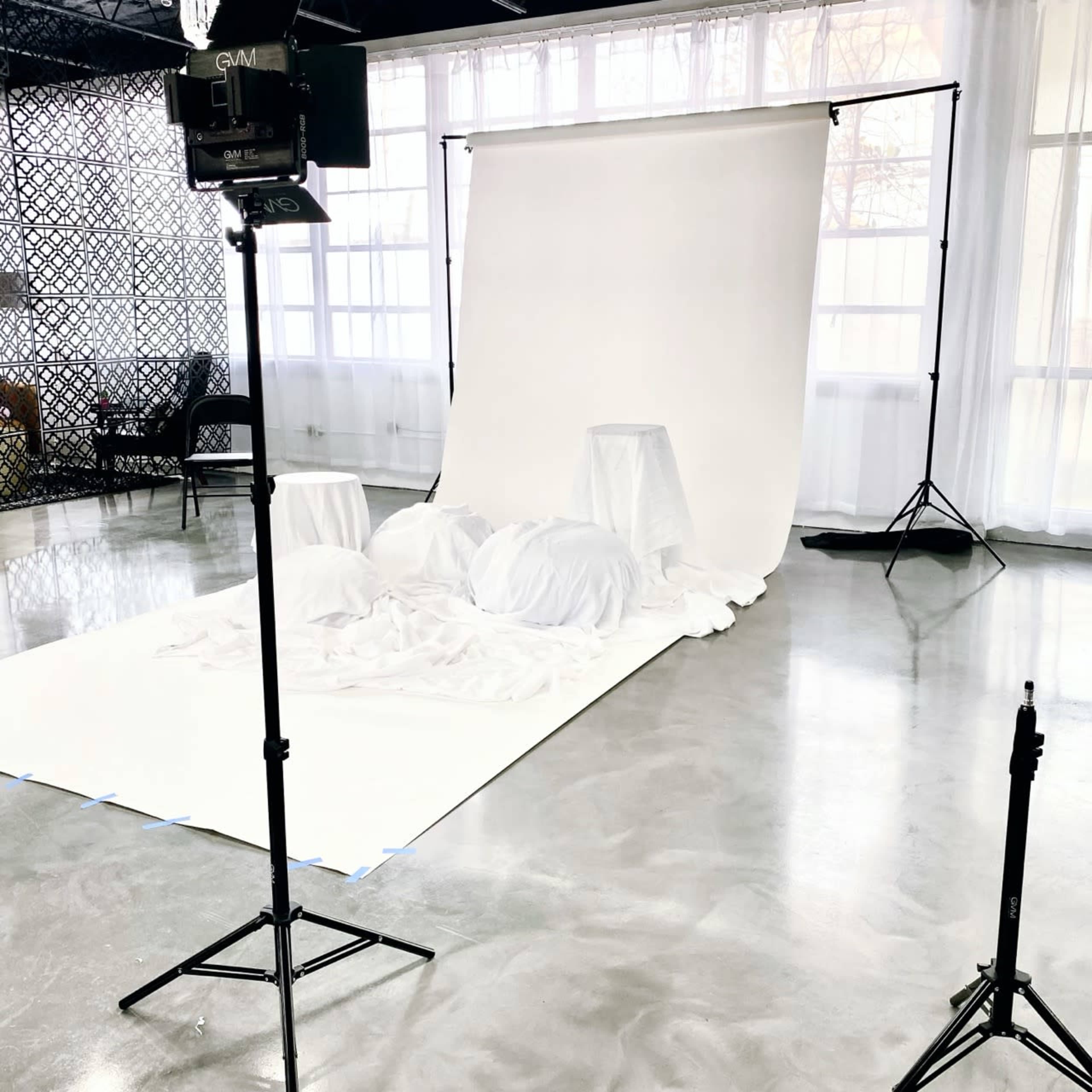 The image shows a photography setup with a white backdrop, a table, and draped white fabric in a spacious studio with large windows.