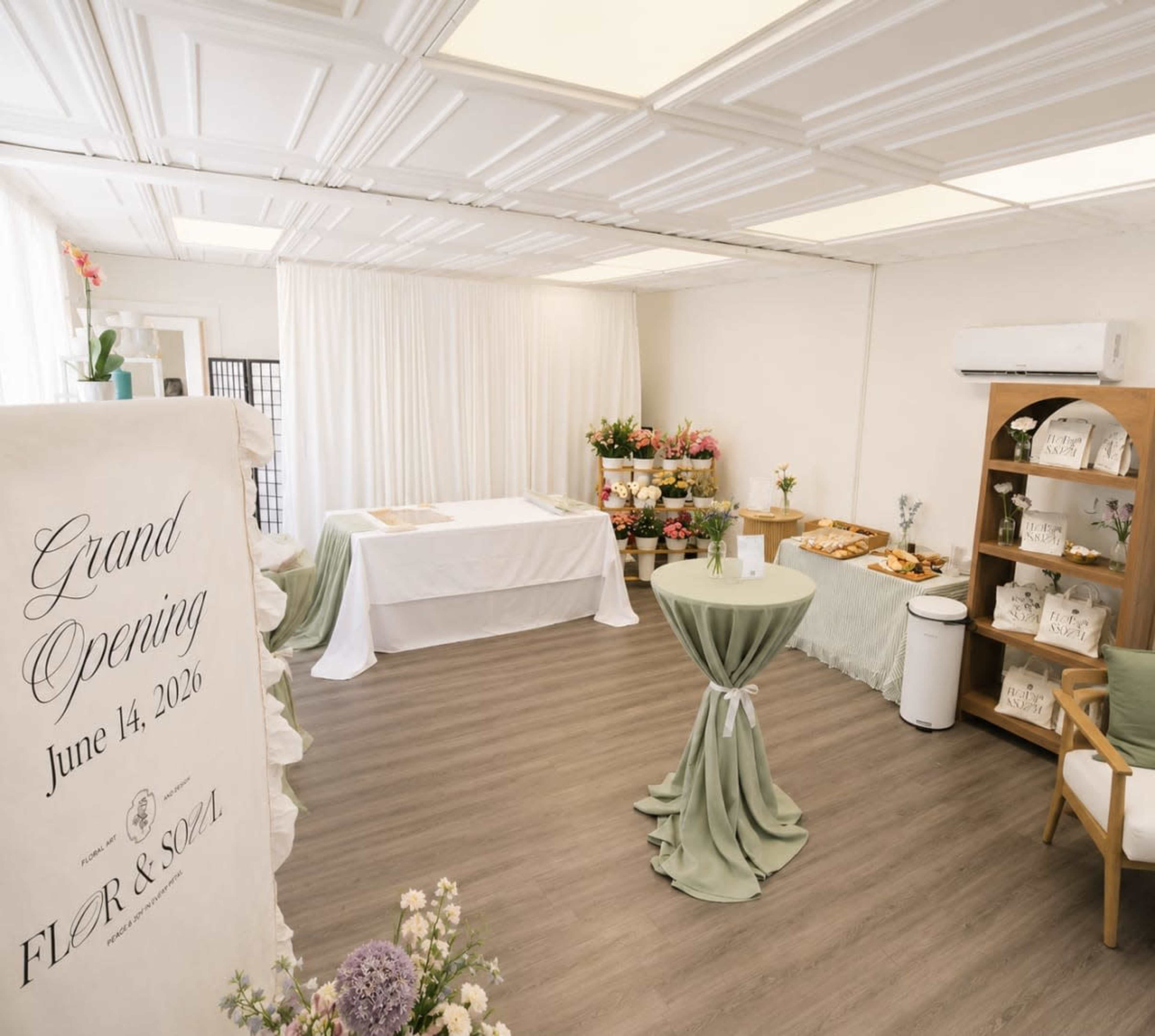 The image shows an indoor event space prepared for a grand opening, featuring a decorated table, floral arrangements, and refreshments displayed on a side table.