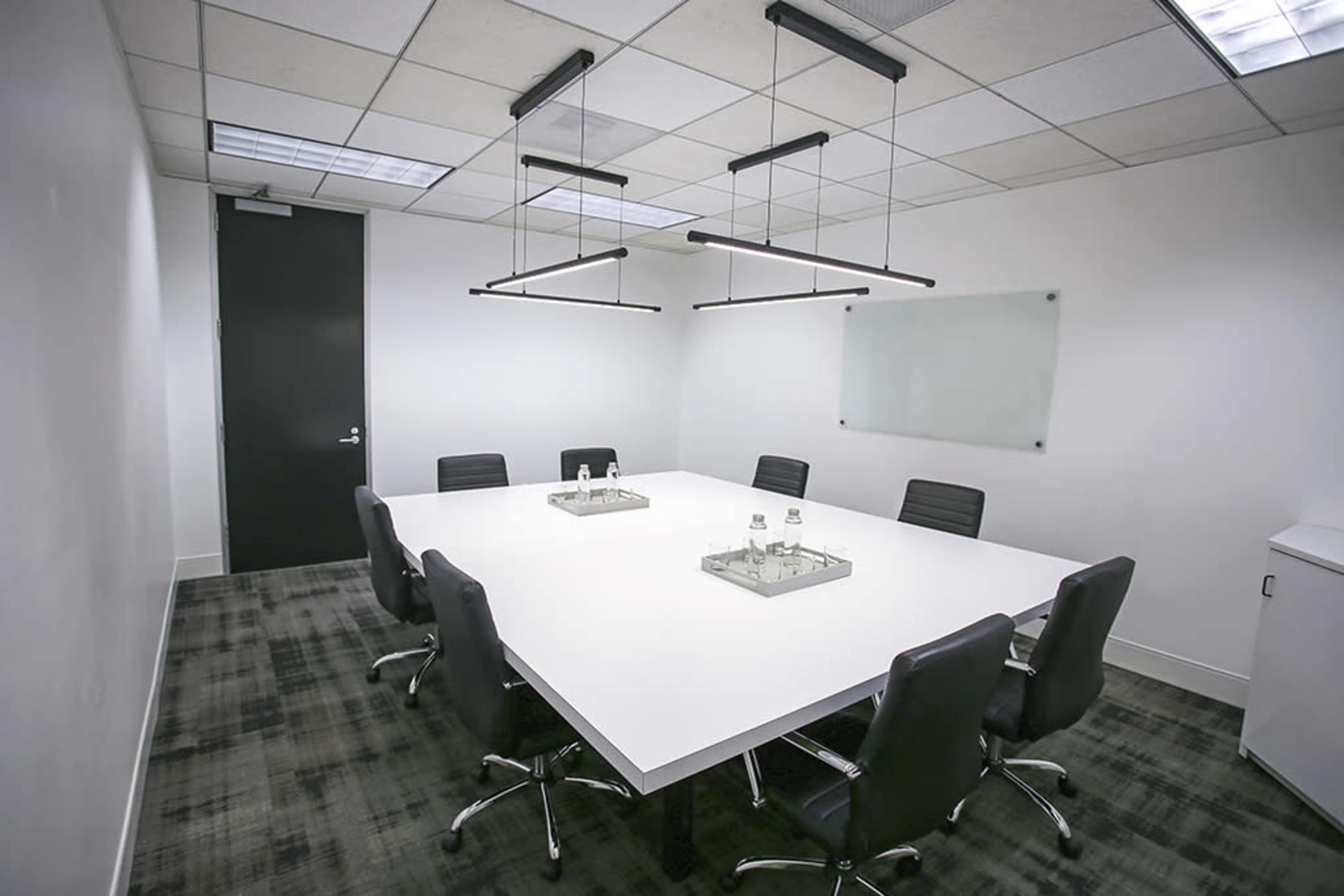 A minimalist conference room features a large white table surrounded by six black chairs under modern overhead lights.