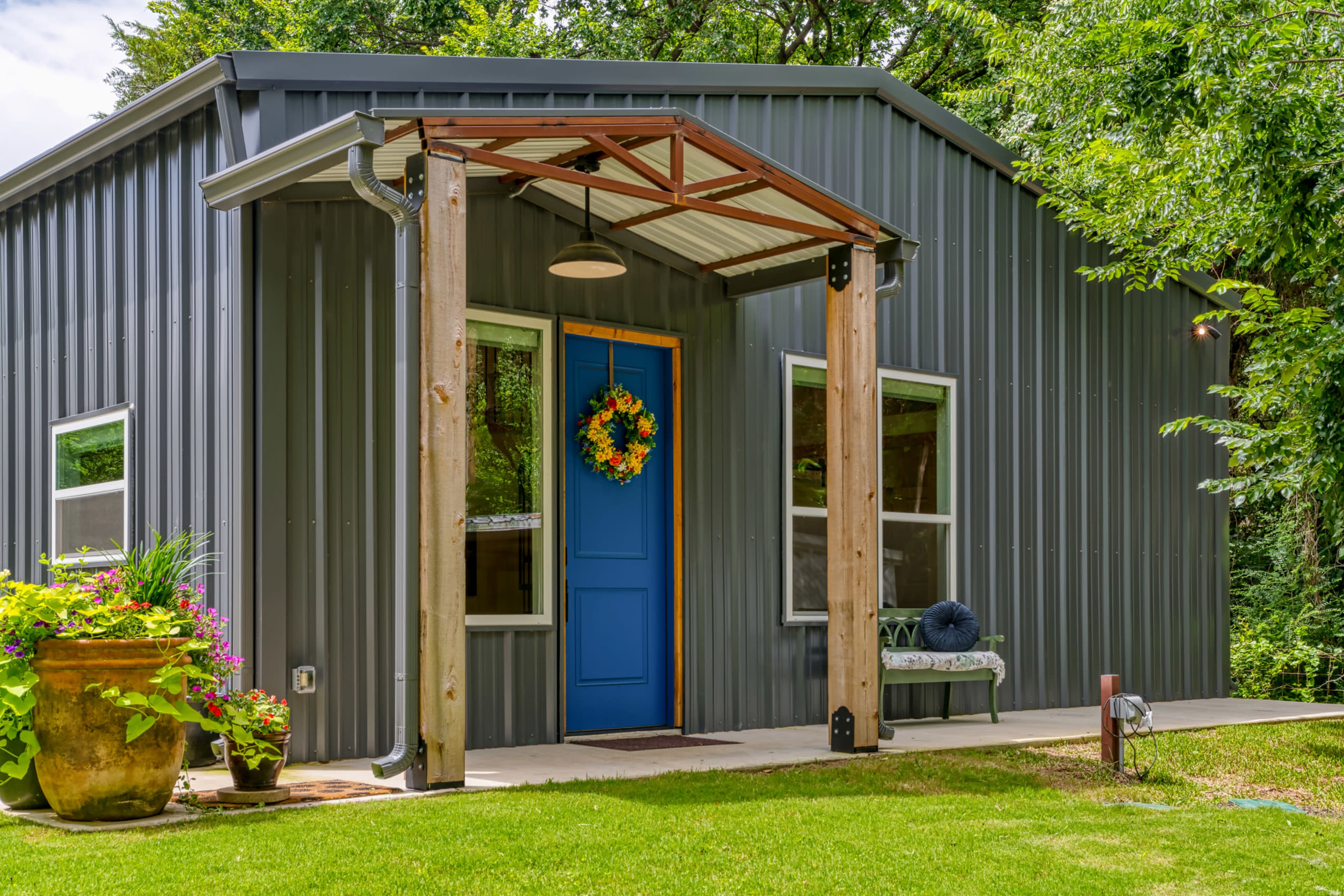 The image shows a modern metal house with a blue front door, an overhead awning supported by wooden beams, and a decorative wreath on the door, surrounded by greenery and a flower pot.