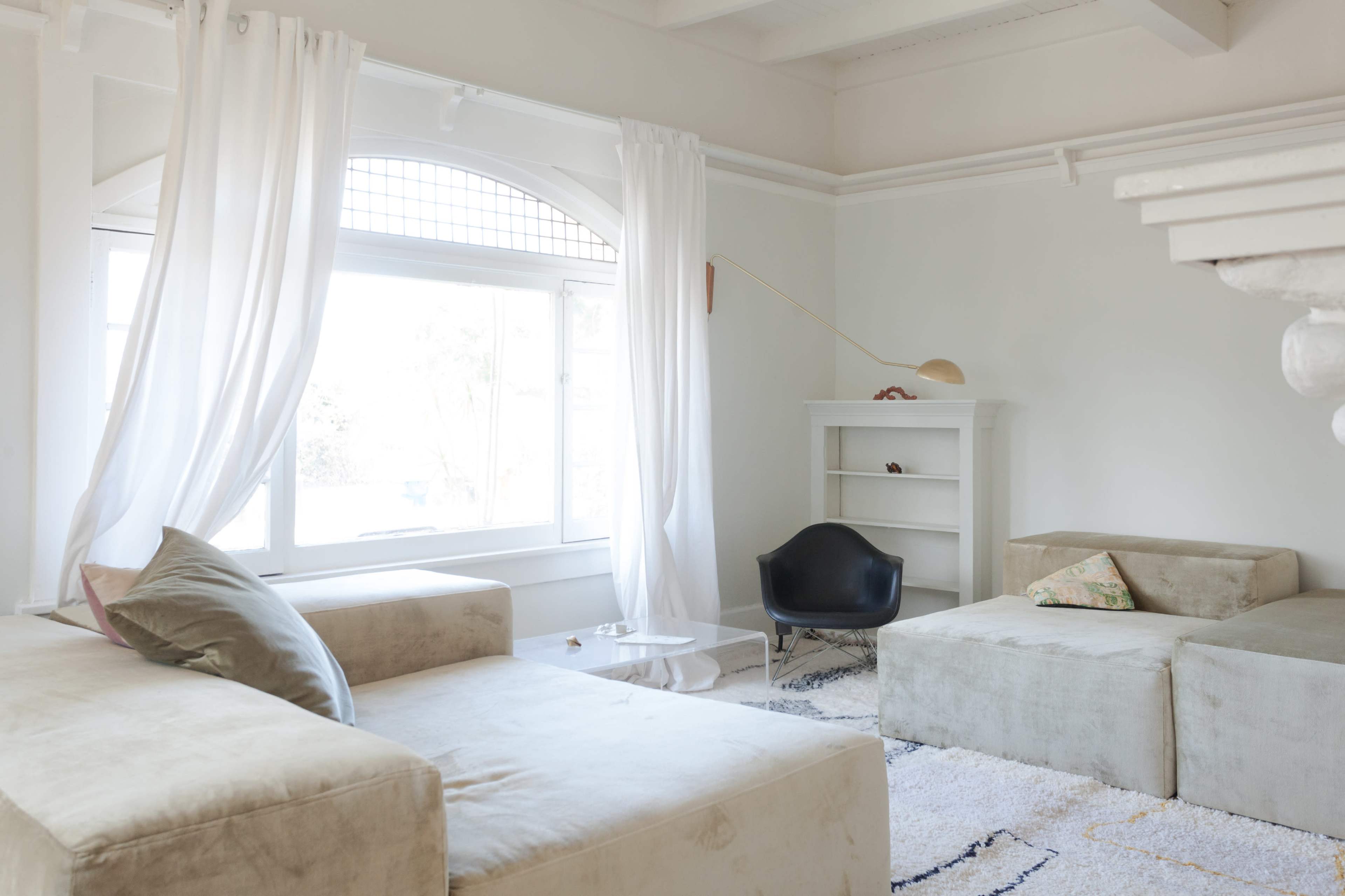 A bright living room features a large window, minimalist furniture, and sheer curtains, creating a spacious atmosphere.