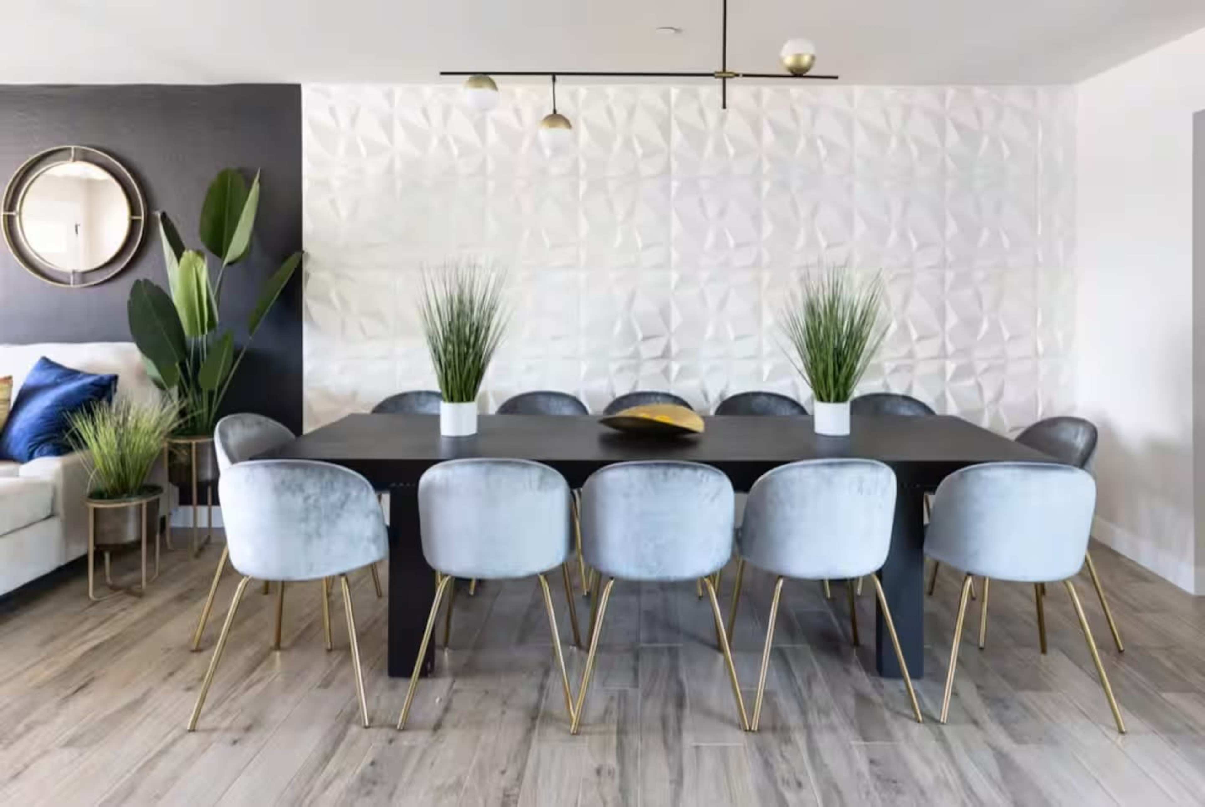 A modern dining room features a long black table surrounded by ten light gray velvet chairs with gold legs, accentuated by decorative plants and a textured white wall.