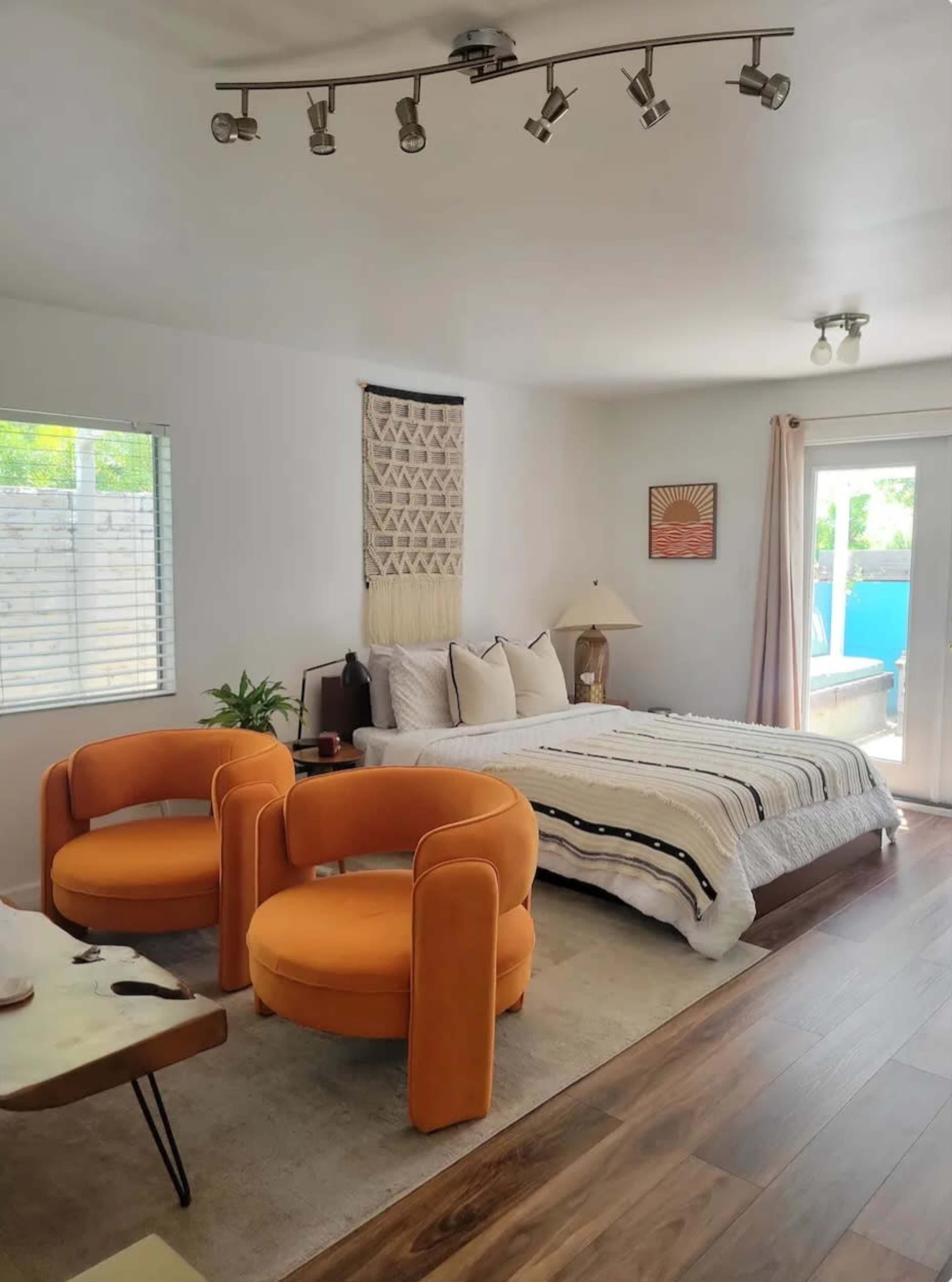 A bedroom features a bed with a patterned blanket, two orange chairs, a table, and a sliding door leading outside.
