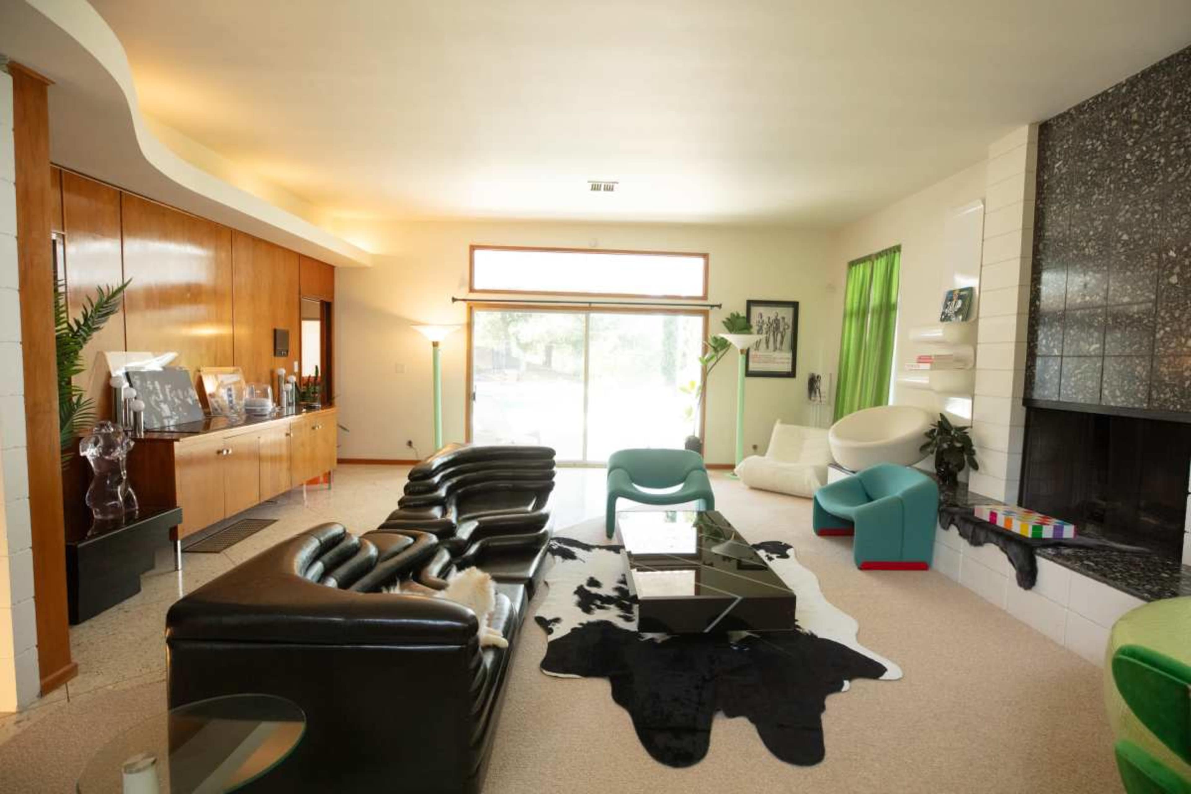 The image shows a spacious living room with a mix of black leather furniture, a white armchair, a large glass door leading to the outside, and decorative elements like a cowhide rug and colorful accents.