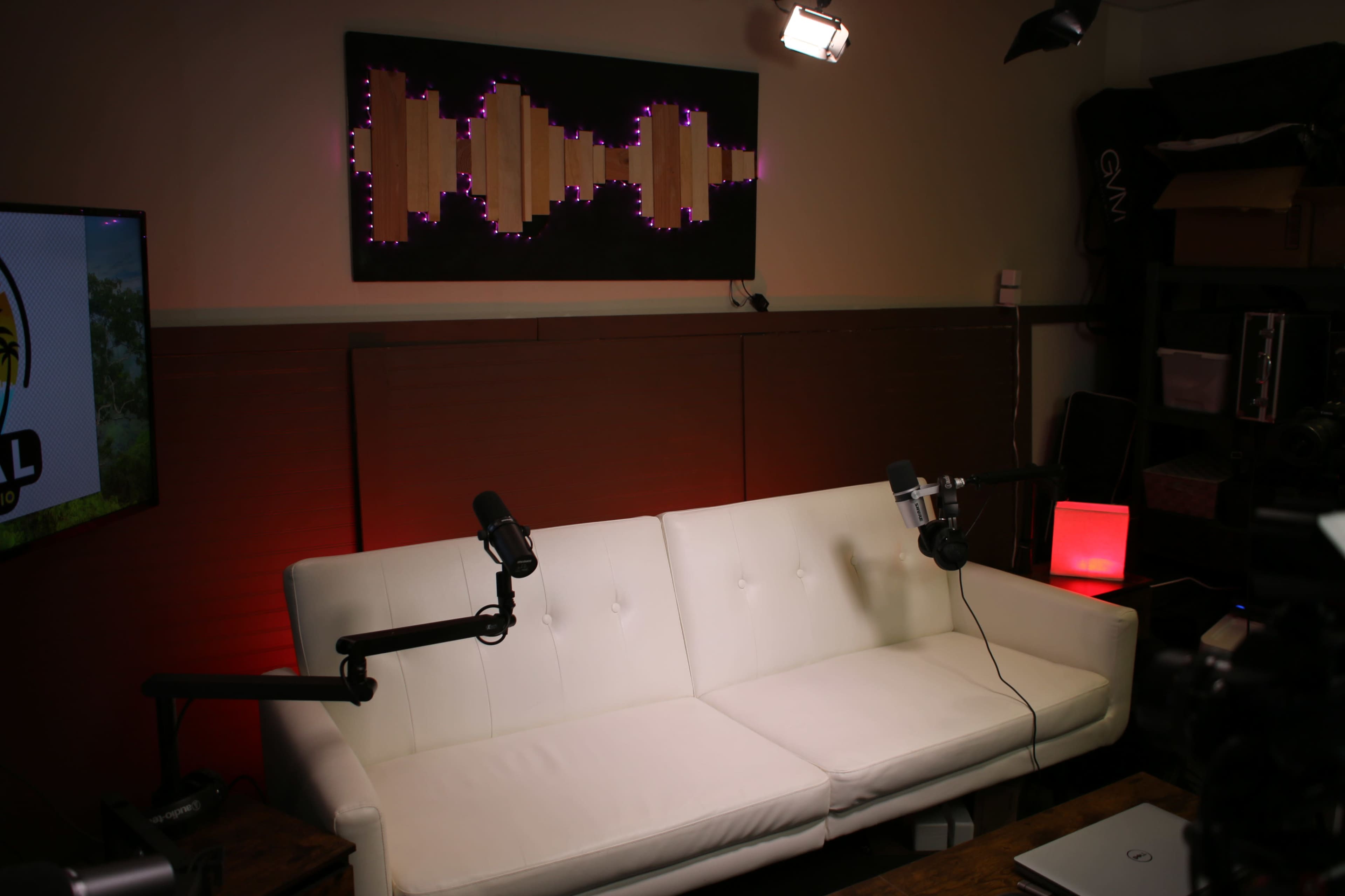 The image shows a modern studio setup with a white couch, two microphones mounted on stands, and a decorative wall art piece illuminated by purple lights.