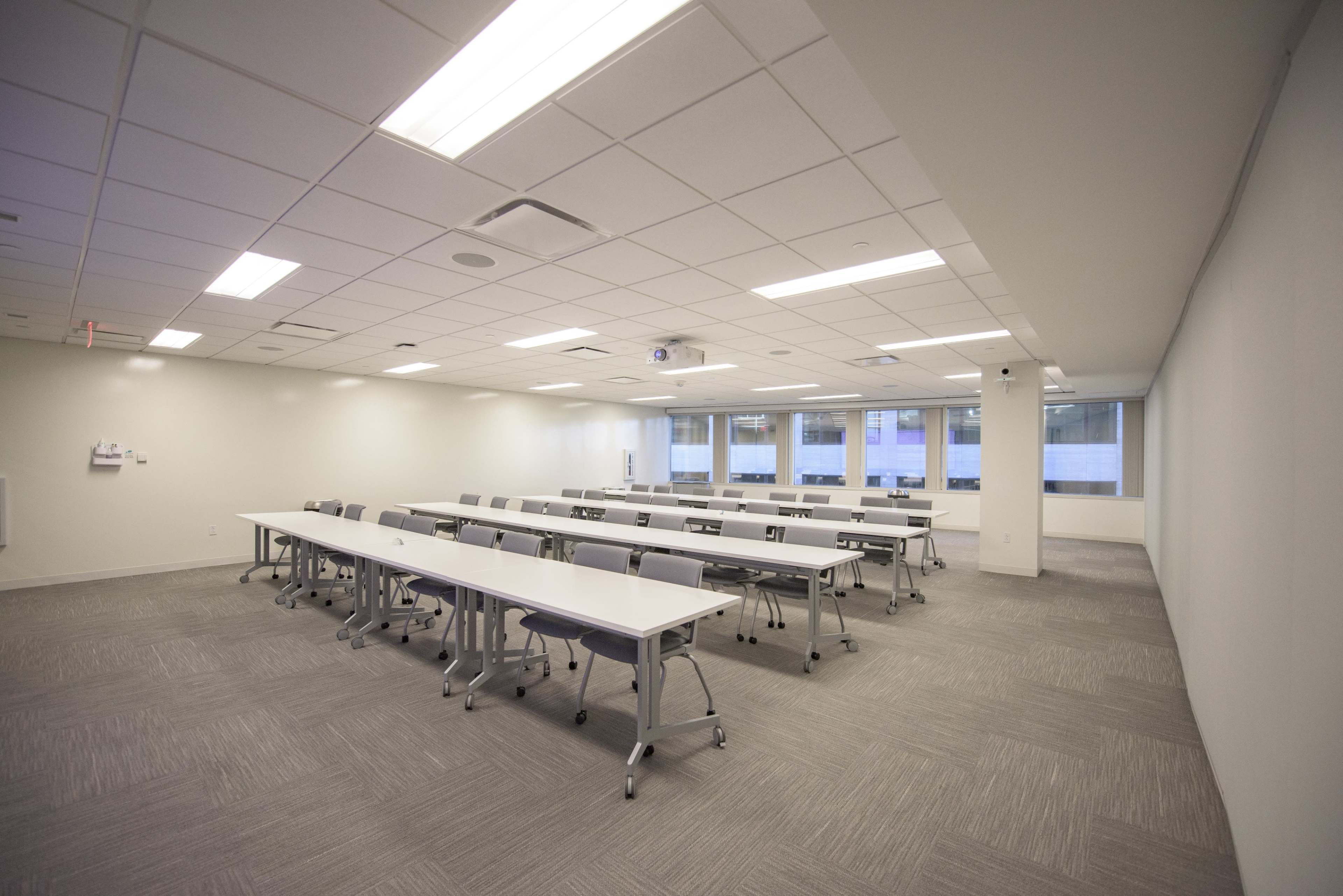 A spacious classroom features rows of long tables with chairs, large windows, and overhead lighting.