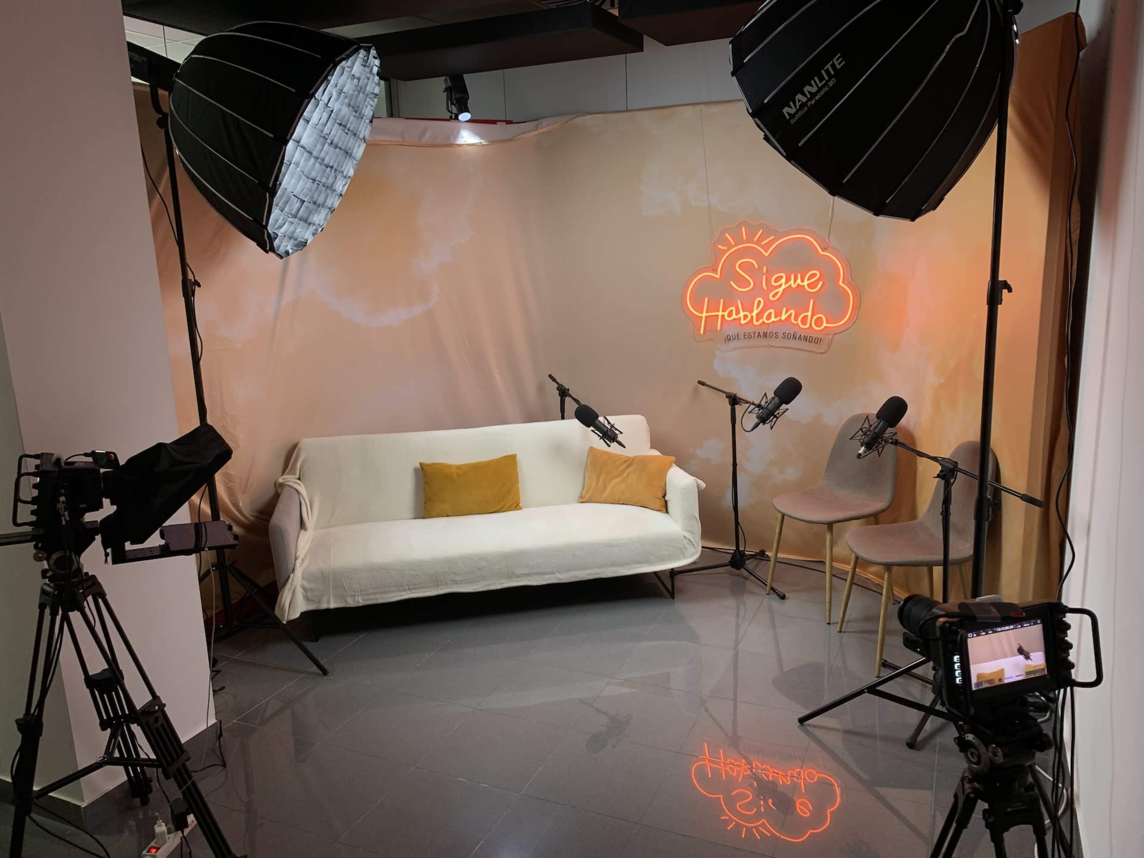 A podcast recording setup featuring a white sofa, a backdrop with the phrase "Sigue Hablando," two microphones, and multiple cameras.