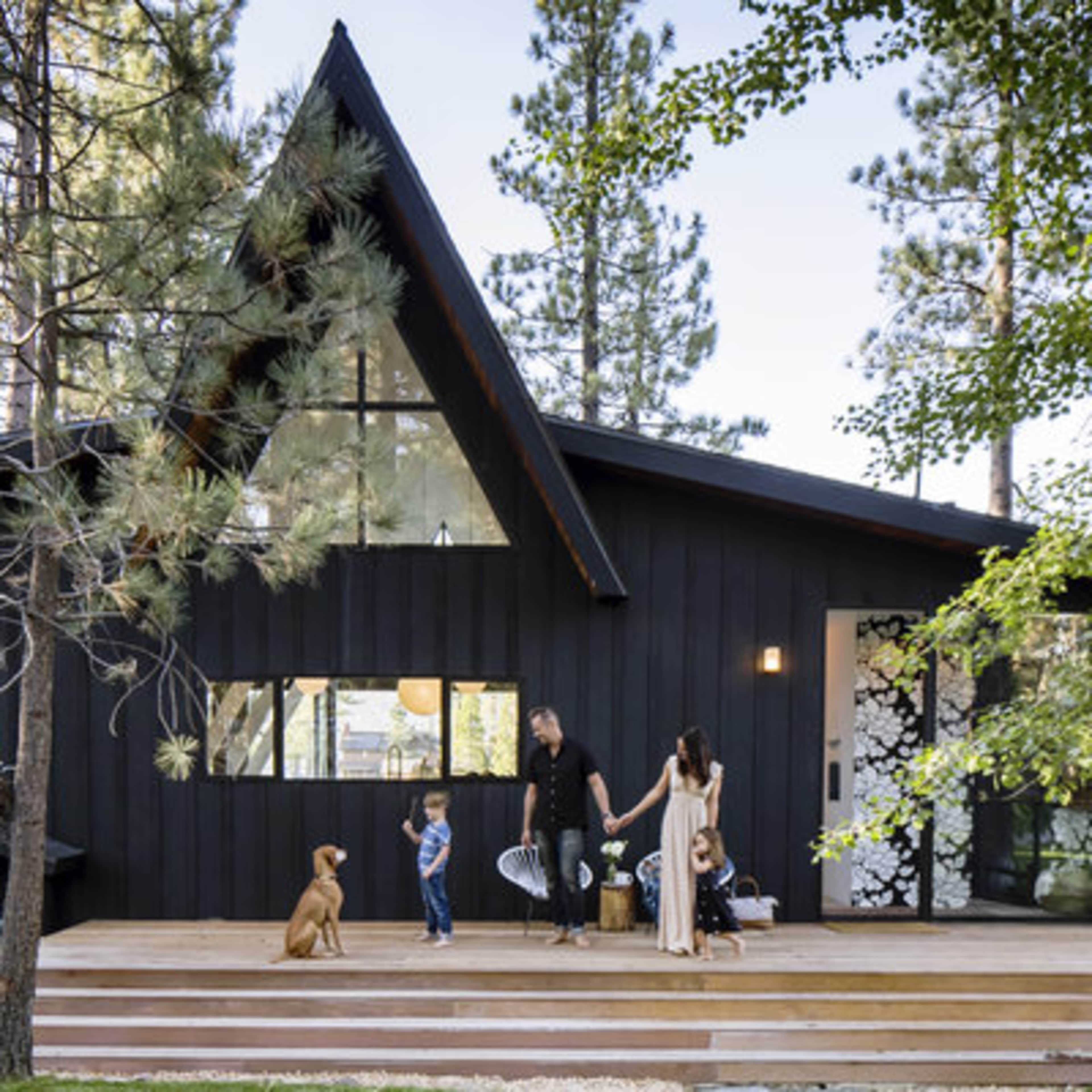 A family with two children and a dog stands on the wooden steps of a modern black house surrounded by tall trees.