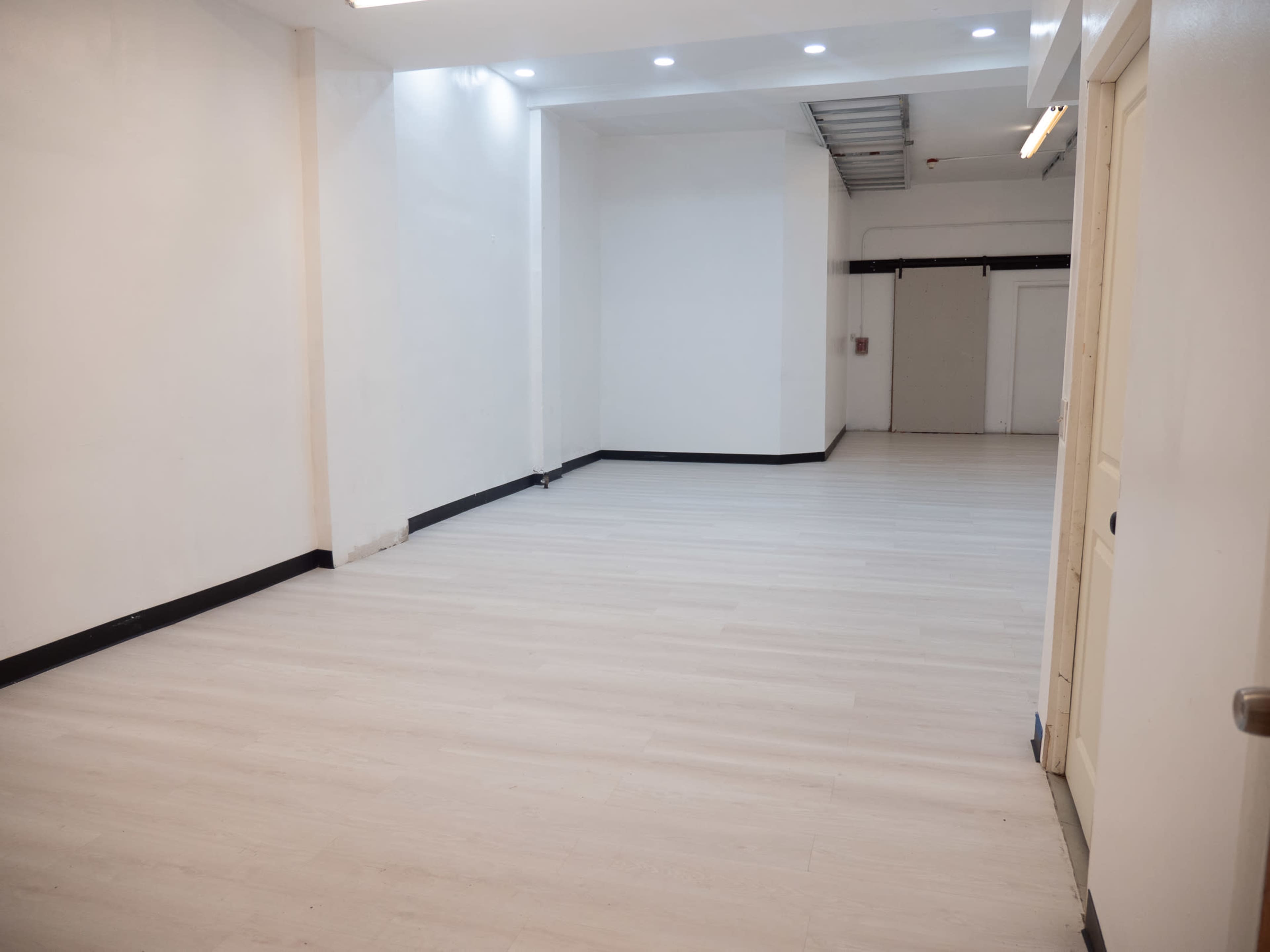 An empty room with light-colored flooring and white walls, featuring a door on the right and a large opening leading to another space at the back.