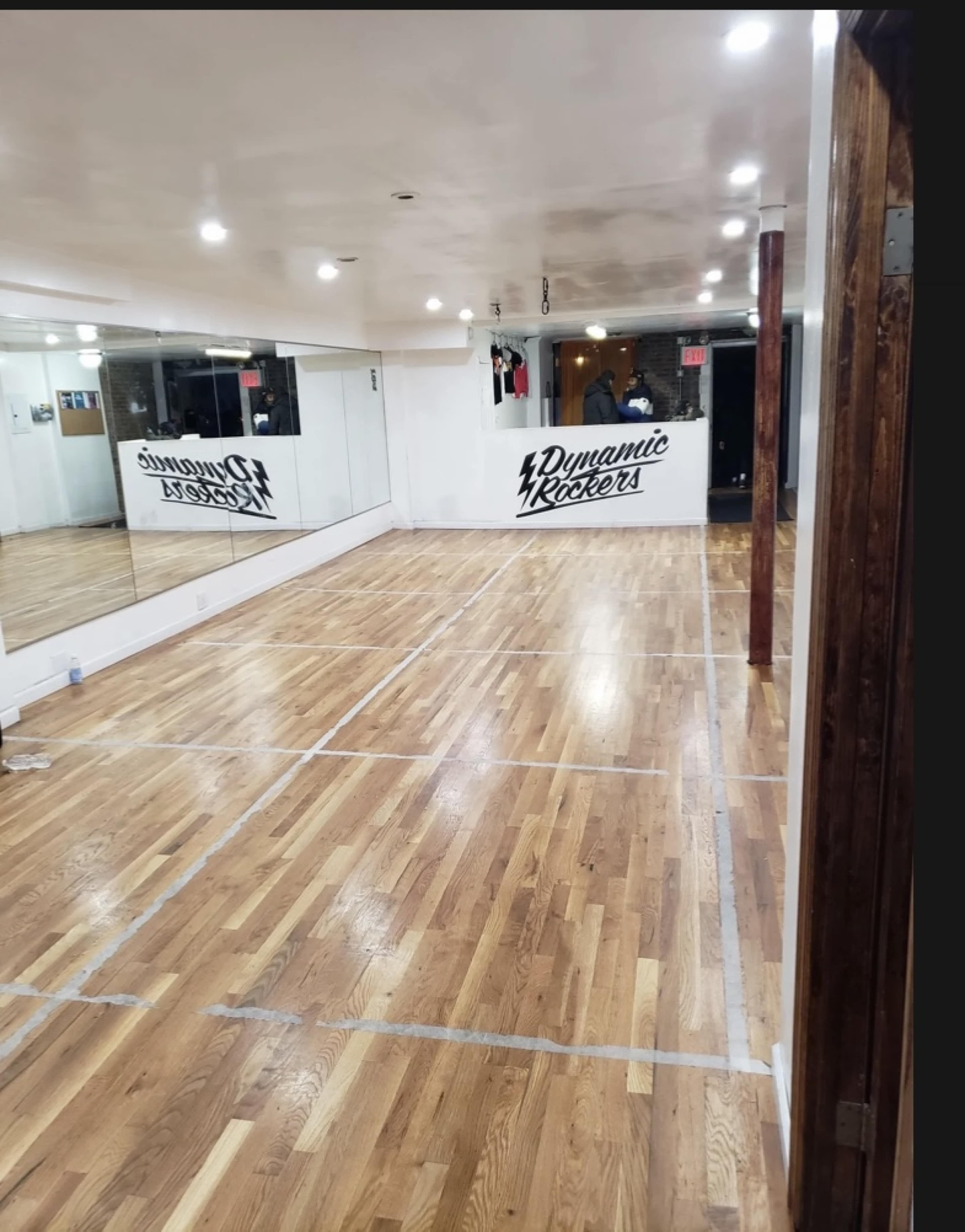An empty dance studio with polished wooden floors and a mirror on one wall, featuring the name "Dynamic Rockers" painted on another wall.