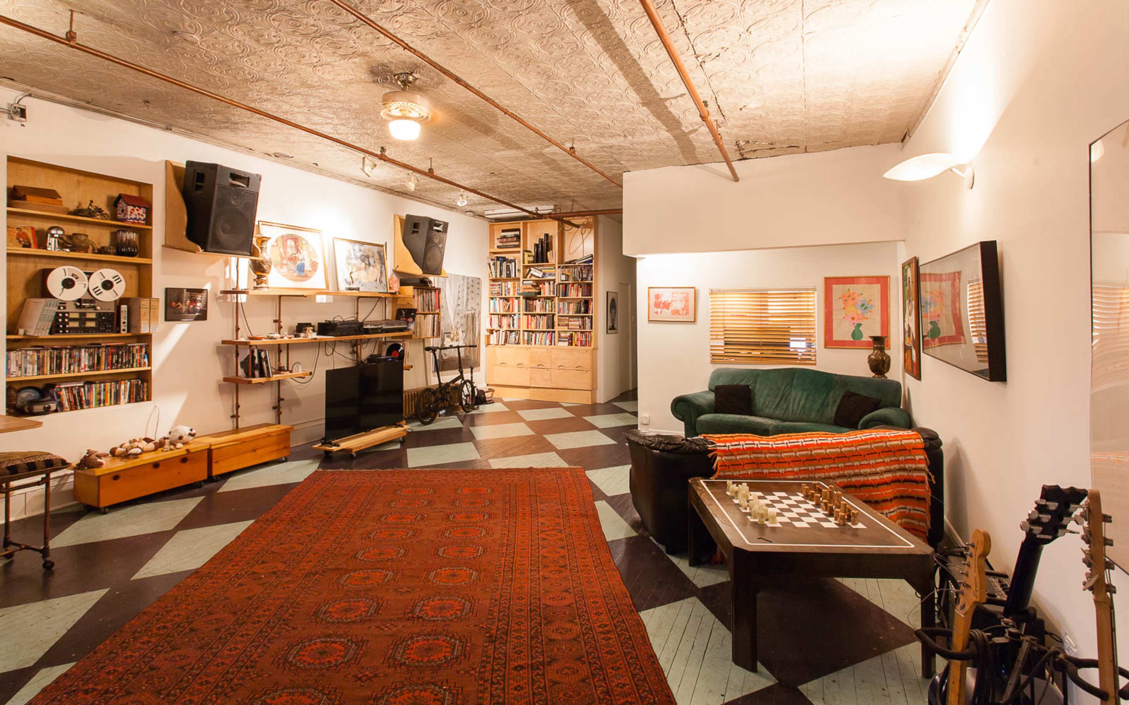A spacious room features a green couch, a chess table, shelves filled with books, large speakers, and various musical instruments against patterned flooring.