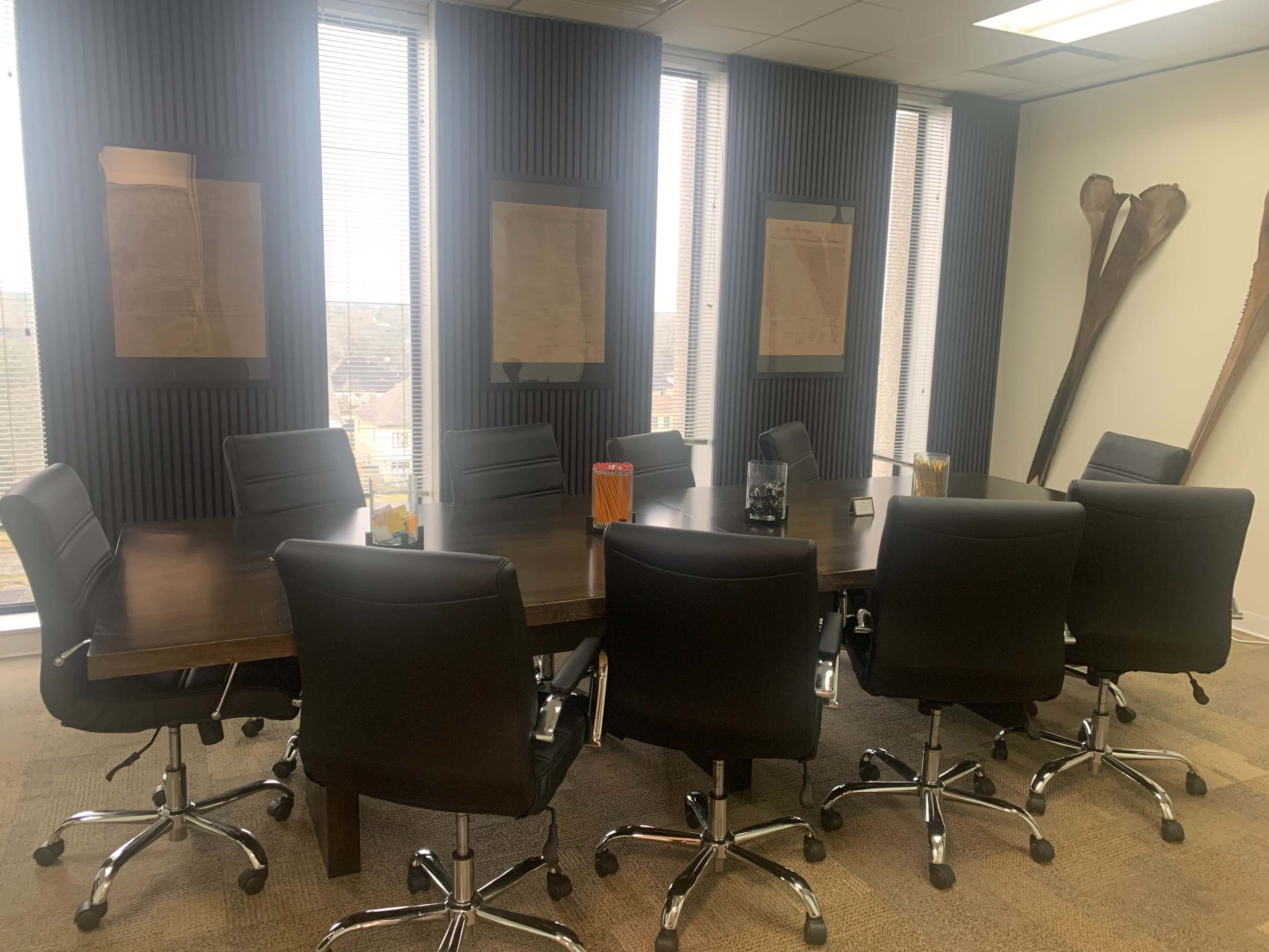 A conference room features a large wooden table surrounded by black leather chairs, with windows on two sides and framed documents on the walls.