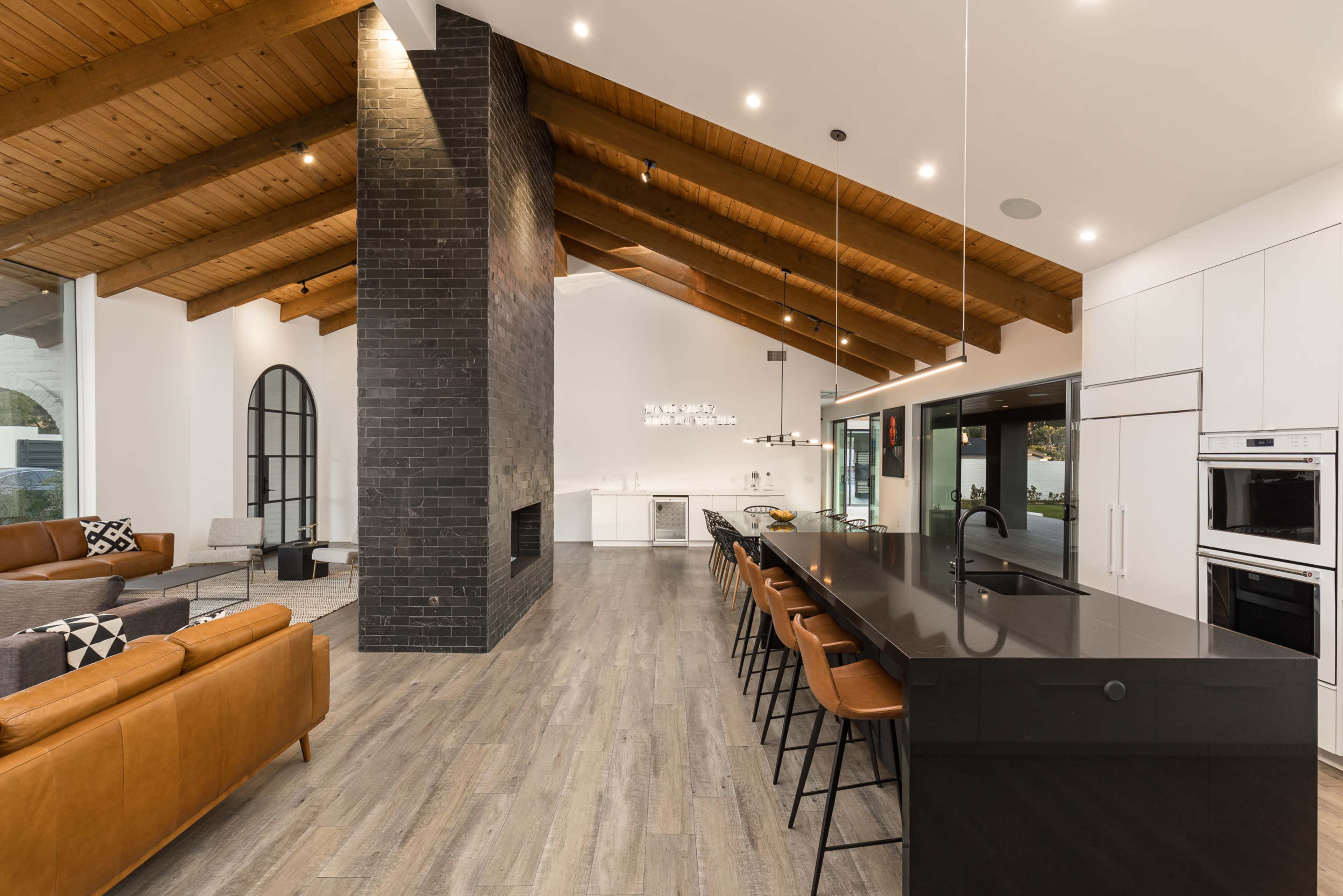 A modern kitchen and living area with a high wooden ceiling, a central black island with bar stools, and a large fireplace set against a black brick wall.