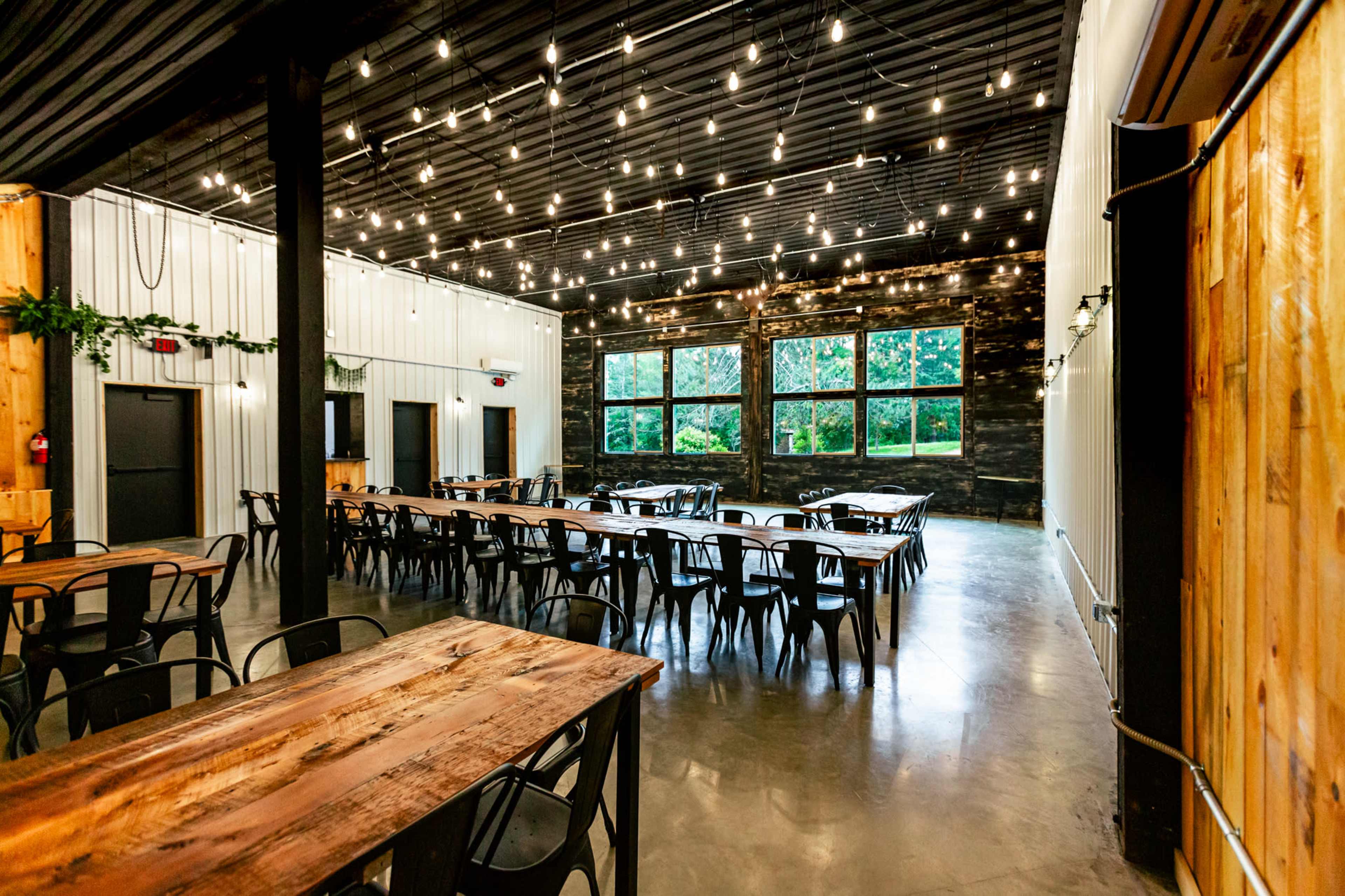 The image shows a spacious indoor venue with wooden tables, black metal chairs, and large windows allowing natural light to enter, adorned with string lights overhead.