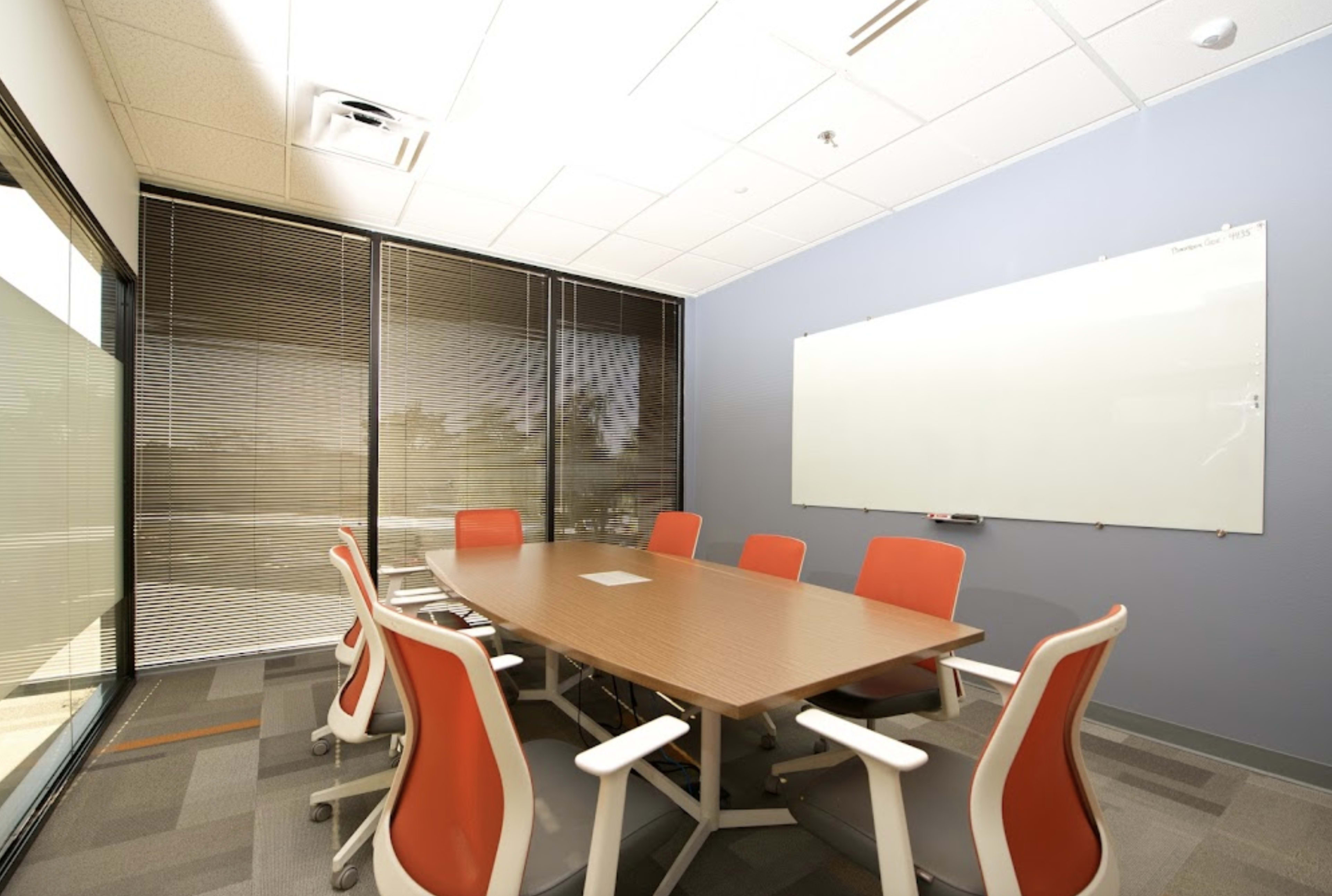 The image shows a modern conference room with a long wooden table surrounded by orange chairs, a whiteboard on the wall, and large windows with blinds.
