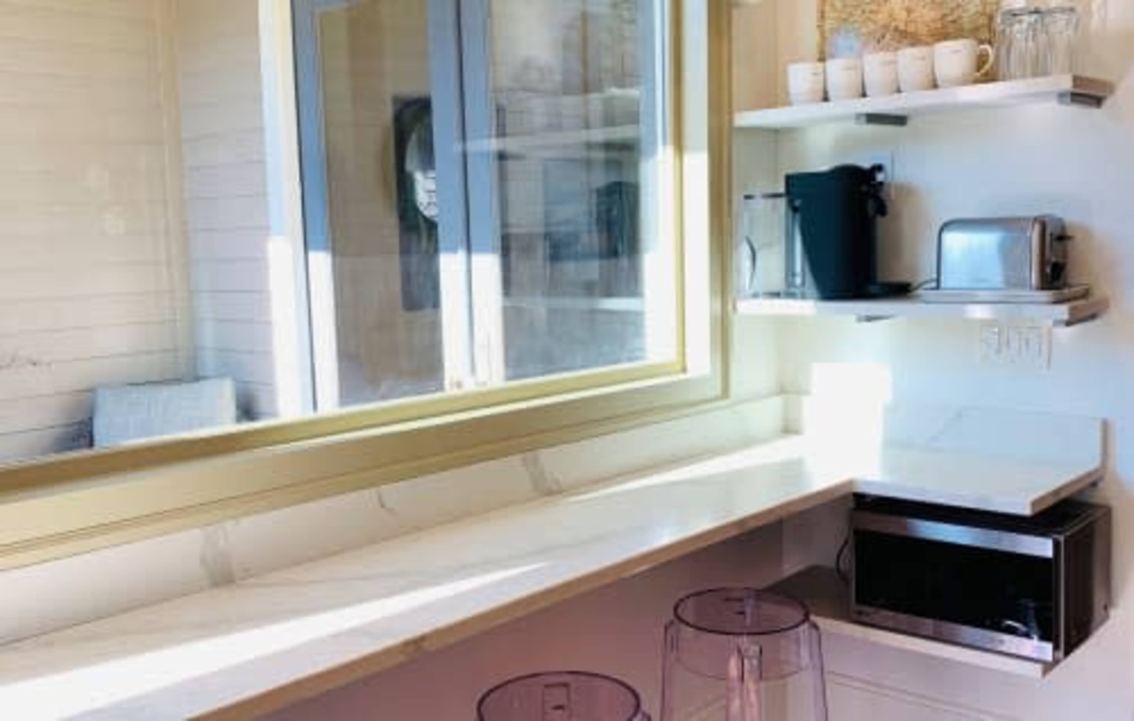 A modern kitchen counter with a window, a coffee maker, a toaster, a microwave, and two transparent stools.