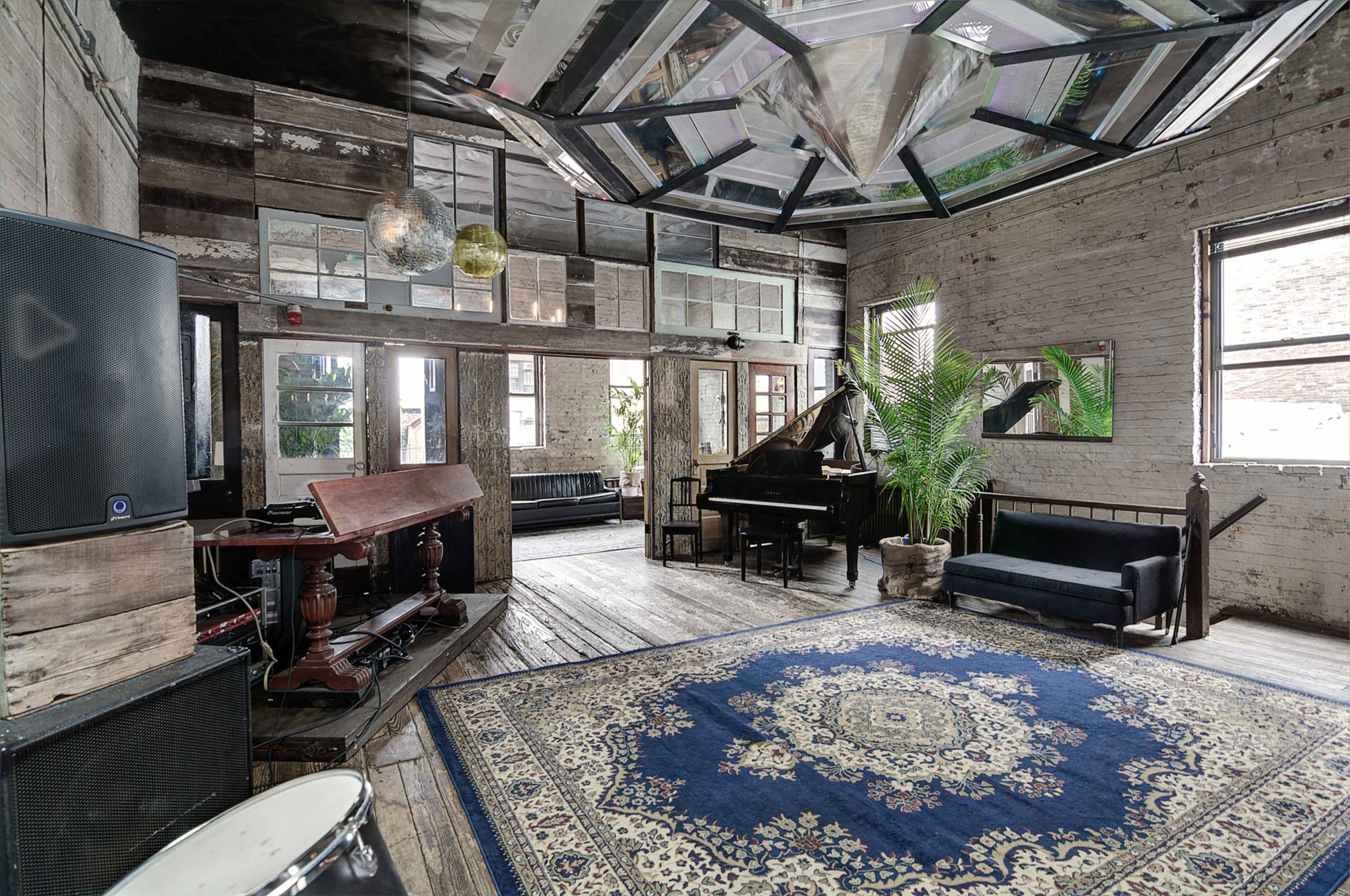 A spacious room with wooden flooring, large windows, and a grand piano, featuring eclectic decor including a large rug and a palm plant.
