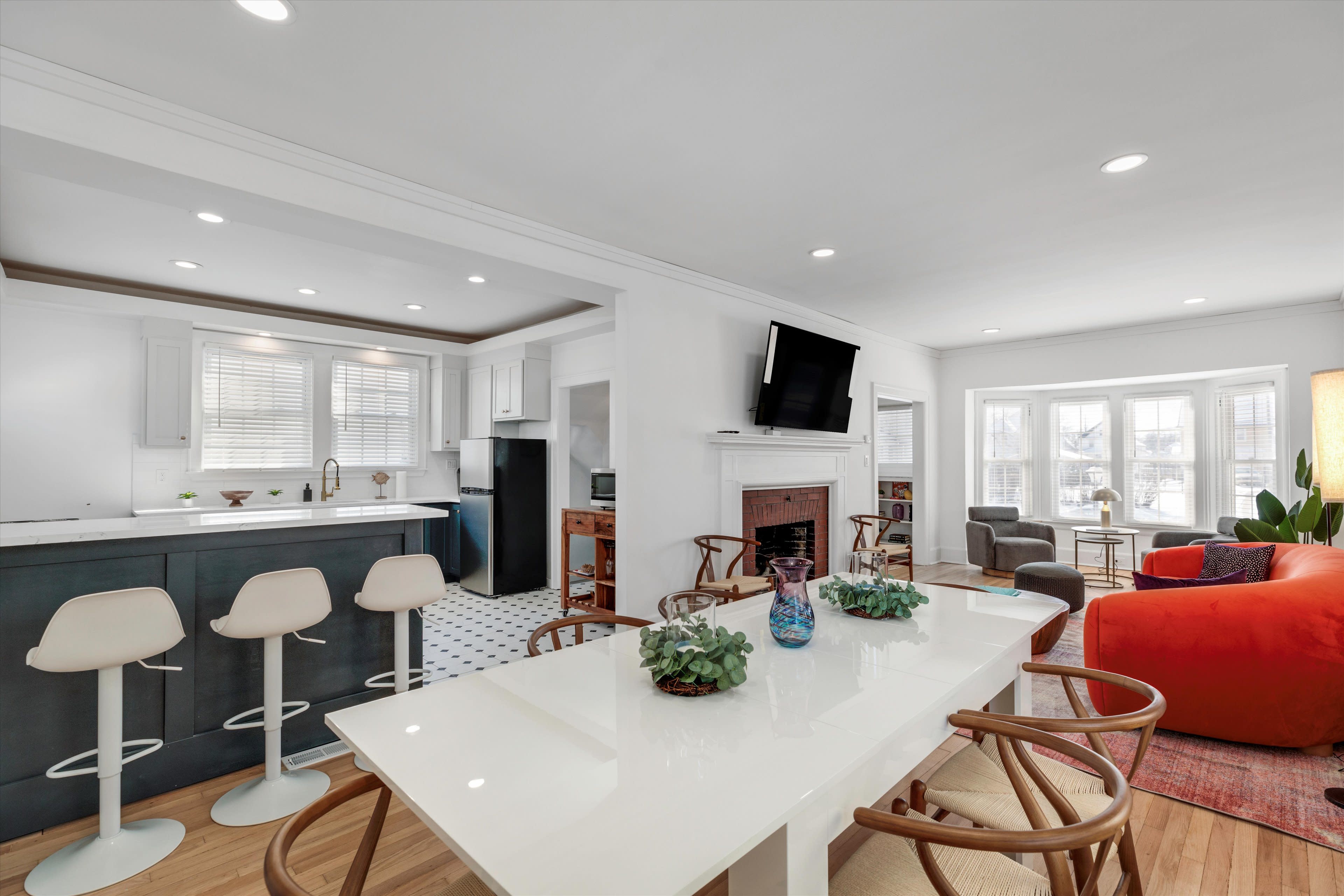 A modern kitchen and dining area features a white table, bar stools, and a living room with a large window and colorful furniture.