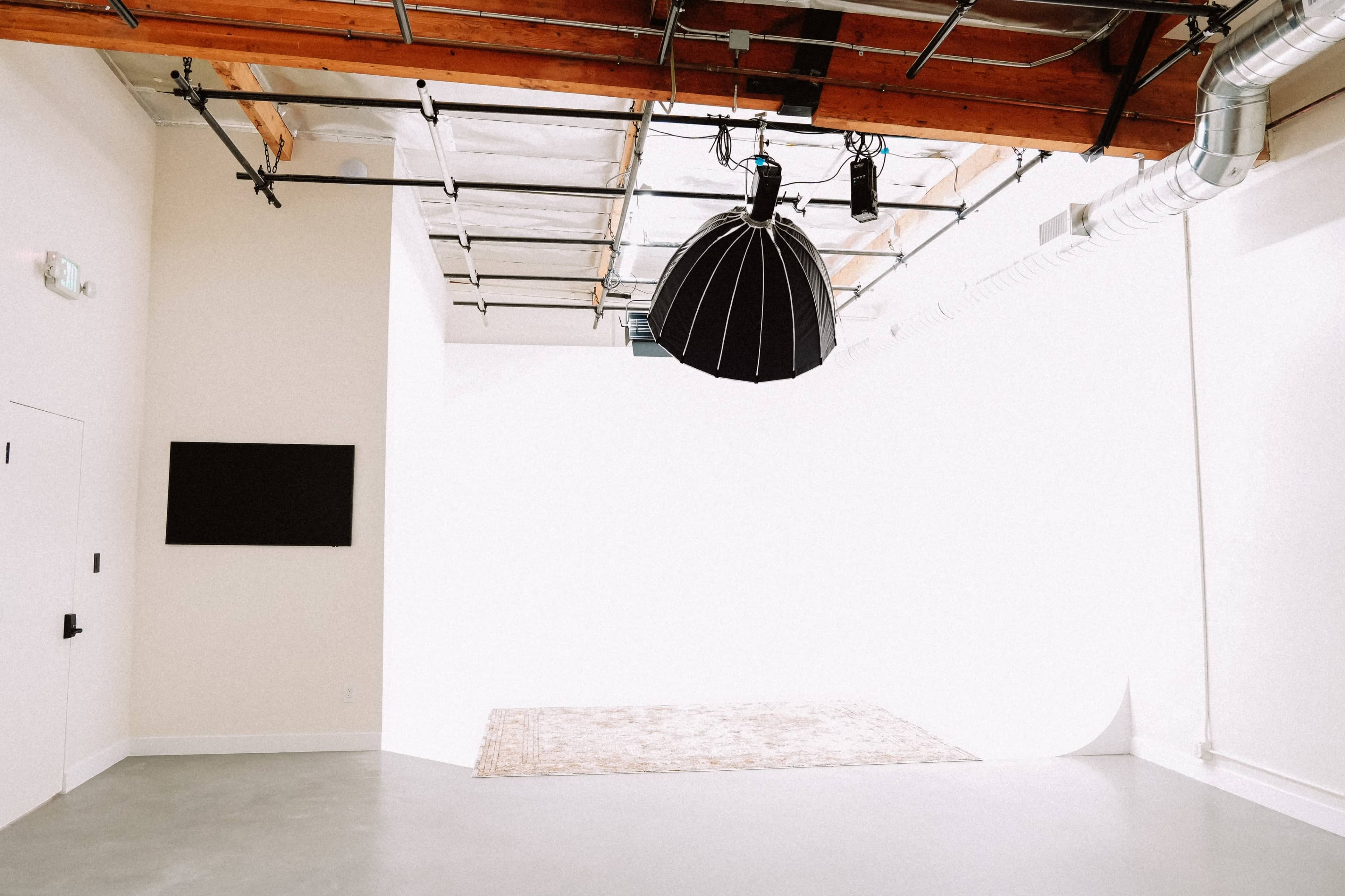 The image shows a spacious, brightly lit room with a large light fixture hanging from the ceiling, a wall-mounted screen, and a patterned rug on the floor.