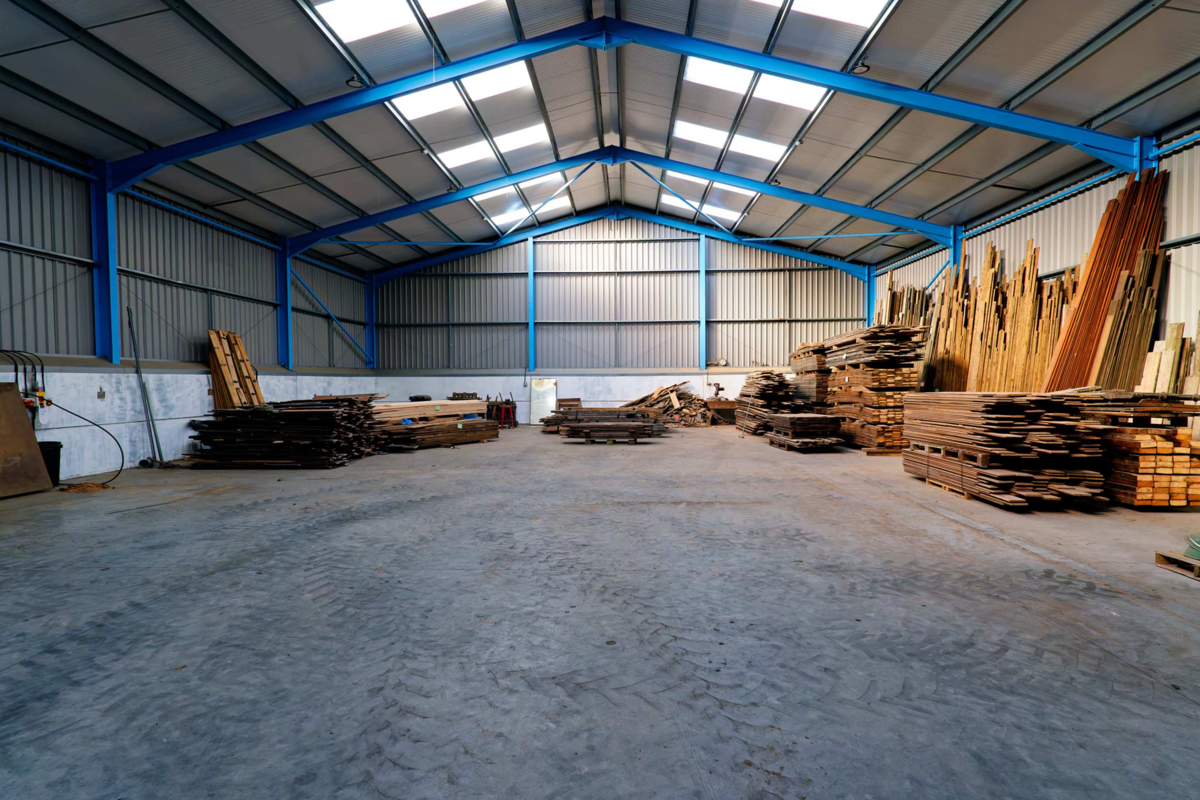 The image shows a spacious warehouse with stacked wooden planks and a concrete floor under a high metal roof.